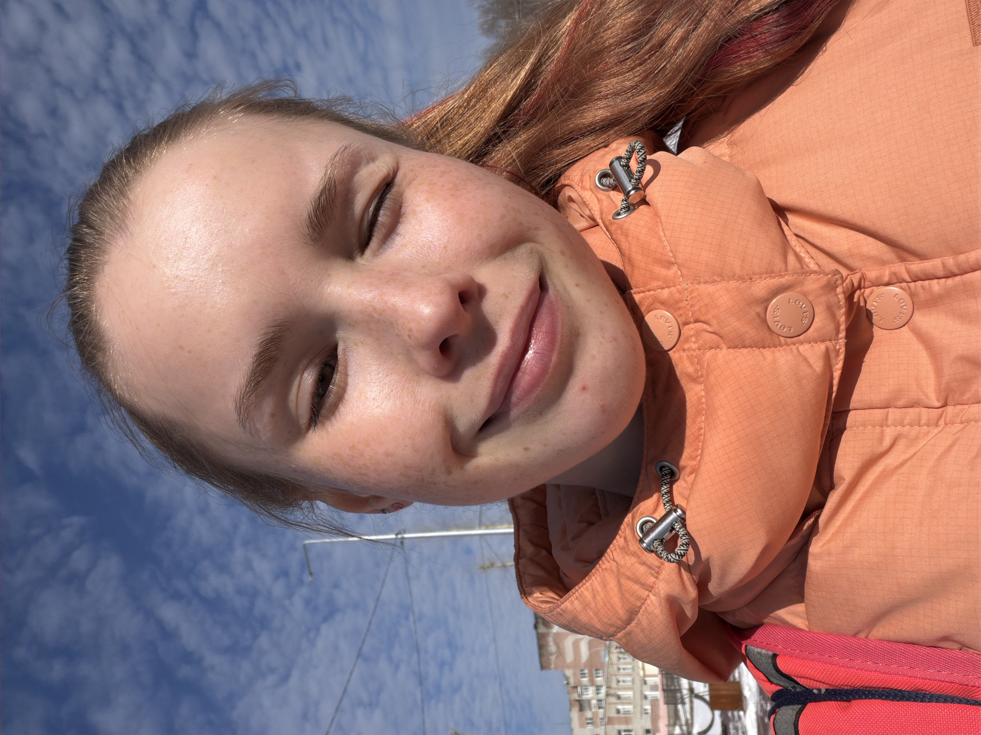 A close-up, rotated portrait features a young female subject with light skin and numerous freckles across her nose and cheeks. She has long, reddish-brown hair, some strands showing a red tint, and her eyes are gently closed as she smiles, basking in bright sunlight. She is wearing an orange or peach-colored puffer jacket crafted from a textured, possibly ripstop, material. Notable details on her jacket include metallic toggles with black and white speckled cords, and circular buttons clearly embossed with the text "LEVI'S 1873". A section of a red backpack or bag is visible at the bottom right.
The background is dominated by a clear, bright blue sky with scattered white cumulus clouds, indicating excellent weather conditions. In the blurred distance, the outlines of multi-story residential buildings are discernible, characteristic of an urban environment. Overhead wires or cables are also visible against the sky. The time of day is bright daylight, suggesting midday or early afternoon. The subject's expression conveys happiness, contentment, and a relaxed enjoyment of the sunny outdoors. The location context for this scene is Berezniki, Russia.