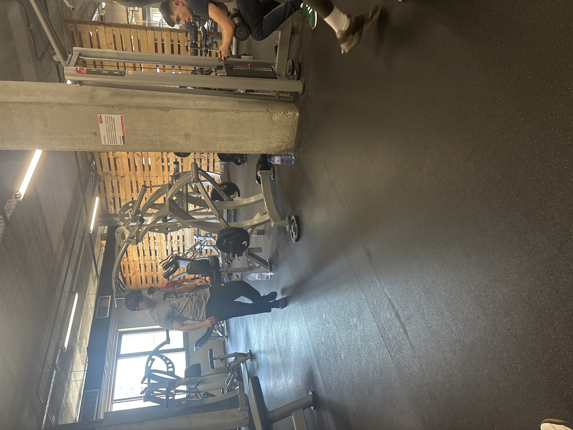 The image depicts a gym interior, showcasing various weightlifting machines. Two individuals are present. One is using a cable machine in the upper portion of the frame, while the other is near a chest press machine in the lower part. A concrete pillar is centrally located, dividing the gym space visually. Pallet-like wooden structures are visible on the upper walls, indicating the building's interior design. Gym equipment such as barbells and plate-loaded machines are sparsely arranged around the central area. A water bottle is seen lying on the floor near one of the machines. Natural light floods the gym through windows. The specific location within Sofia, Bulgaria cannot be determined based solely on the image.