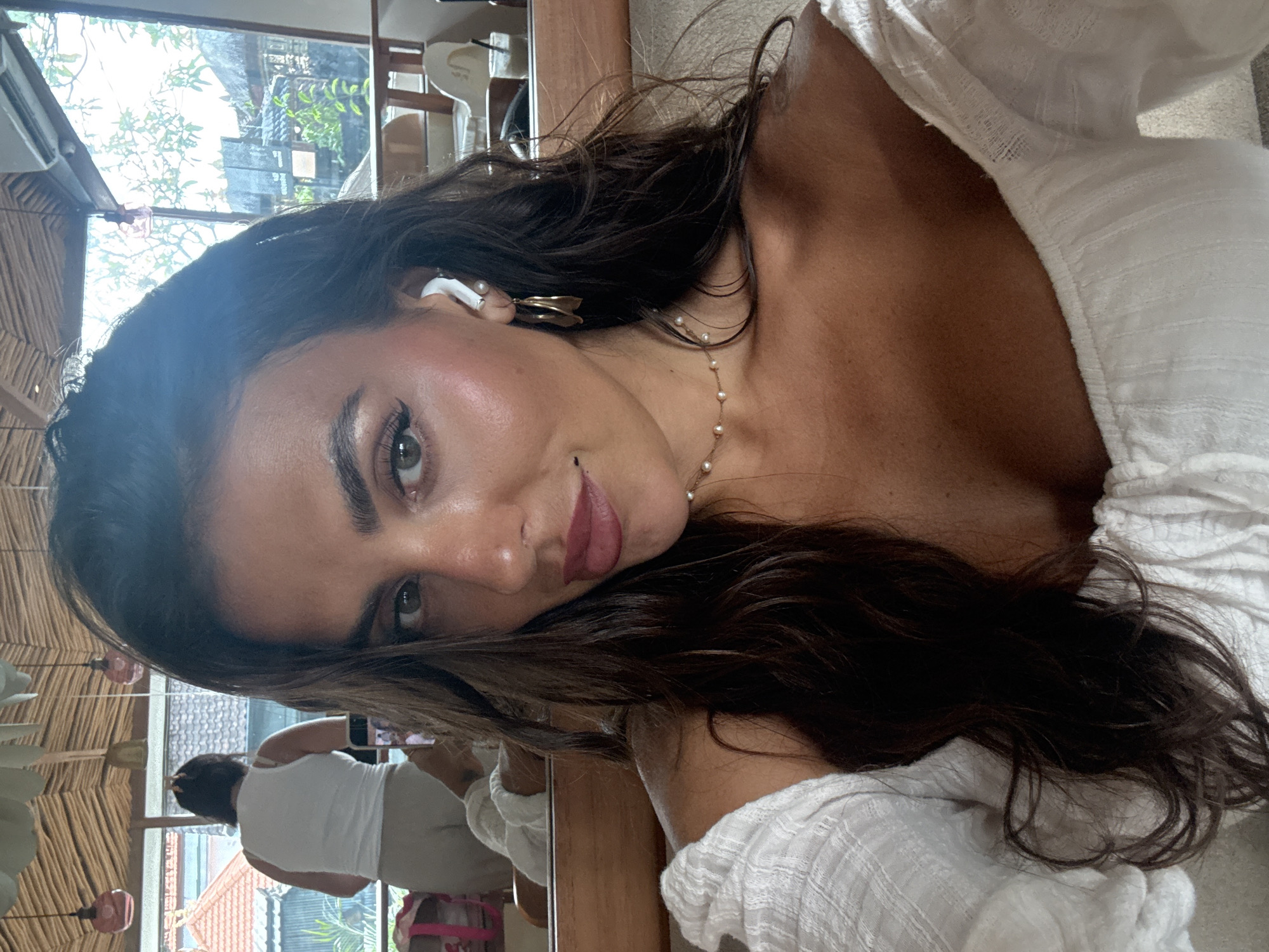 A woman is seen in a selfie-style photograph taken indoors. The subject has long, dark hair and is wearing a white top. She has an earbud in one ear and is wearing a gold-colored earring. A pearl necklace adorns her neck.
In the background, visible through a window, greenery and buildings suggest an outdoor environment. A reflection of a person in a white top and light-colored pants, possibly taking a photo, is visible in a mirror behind the subject.
The location is suggested to be in North Kuta, Indonesia.