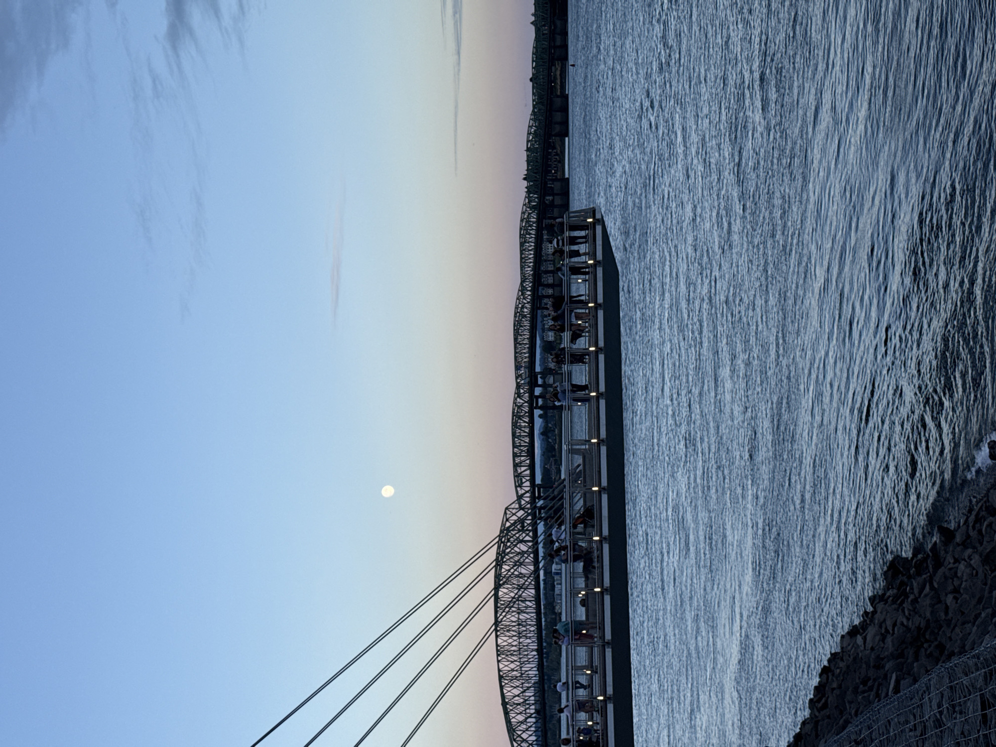 The image depicts a twilight scene at the Vancouver Waterfront in Vancouver, Washington, United States of America. In the foreground, choppy blue water occupies the lower half of the frame, with a rocky shoreline visible on the left. A promenade or pier extends from the left into the water, with several people standing and observing the view. A dog is also present on the promenade. Overhead, thick cables descend from the left, attached to the promenade's structure. In the background, a large, arched bridge spans across the water, with another bridge visible further in the distance. The sky transitions from a soft pink and orange near the horizon to a pale blue overhead, with a bright full moon positioned in the upper left quadrant of the sky. The overall atmosphere is serene and captures the transition from day to night.