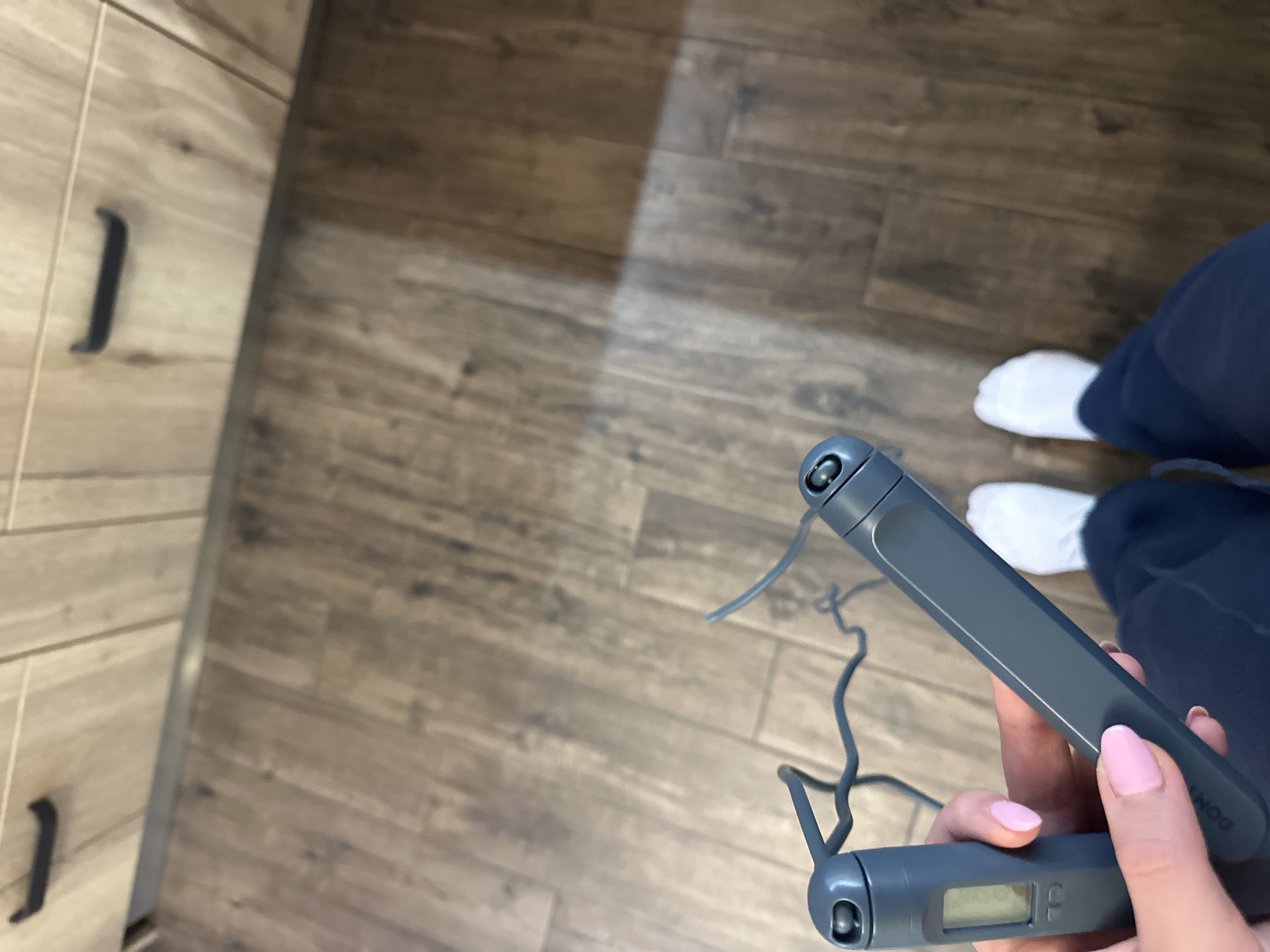 An interior scene is depicted from a first-person, downward perspective. A person's left hand, with pink-manicured fingernails, grasps a grey digital skipping rope. One handle features a small screen display, and the other handle bears the faint inscription "DOMYOS." The dark grey rope is partially visible, coiled between the handles. Below, the person's feet, clad in white socks and dark blue trousers, are positioned on a brown wood-grain laminate floor. In the upper background, light-colored wooden cabinetry with dark horizontal handles extends above a metallic kick plate. A distinct rectangular reflection of light is present on the floor. The specific location within the city of Sofia, Bulgaria, cannot be determined from the visual information provided.