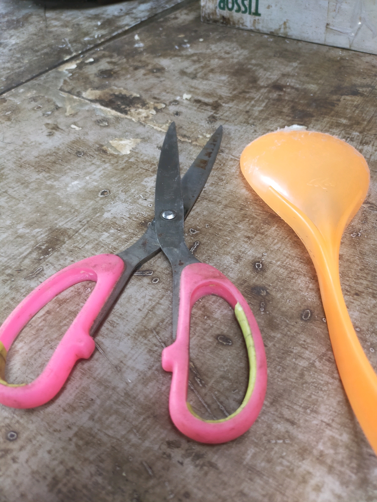 The image shows a close-up, top-down view of a pair of pink-handled scissors and an orange plastic ladle resting on a worn, wooden surface. The wooden surface appears to be a table or countertop, with visible signs of age and use, including scratches, stains, and some white debris. The scissors are open, with their metallic blades positioned towards the top of the frame. The ladle is positioned to the right of the scissors, with its scoop facing upwards. In the background, a portion of a box with text is visible, though it is out of focus. There is no indication of people or any specific actions occurring in the scene. The overall environment appears to be indoors, likely a workshop or a place where tools are kept. The location of Mojodanu, Indonesia is not ascertainable from the visual information provided in the image.