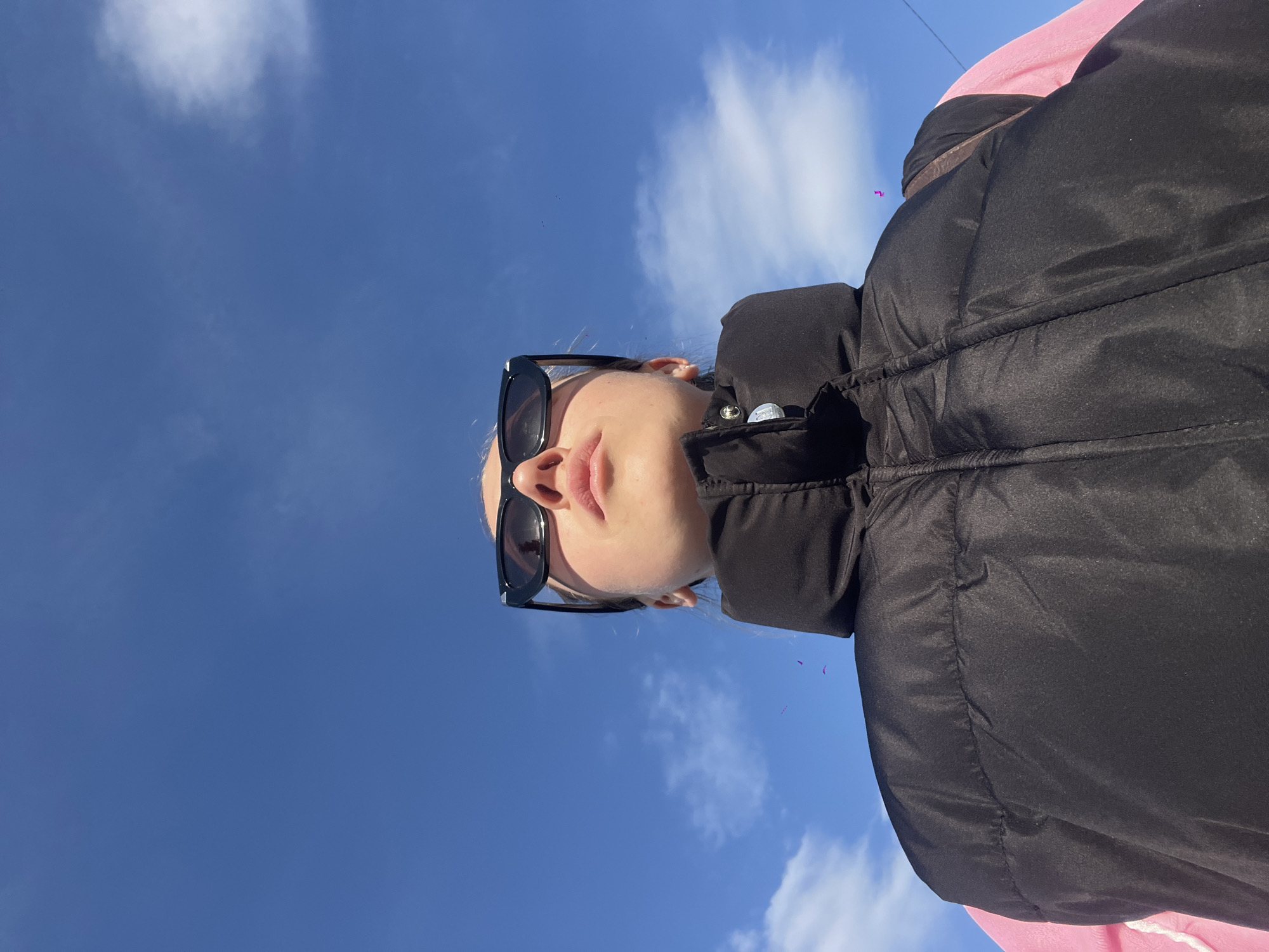 A woman with fair skin, wearing black sunglasses and a black puffer jacket over a pink garment, is positioned in the foreground, looking upwards. The background is a clear, blue sky with scattered white clouds. The location is Boreti, Montenegro.