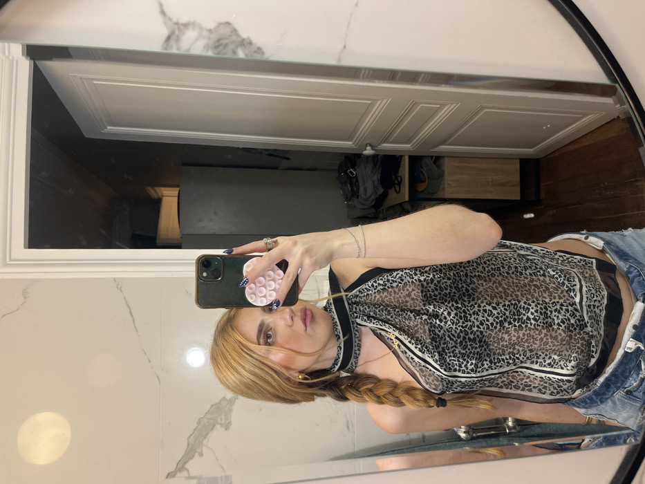 The image captures a mirror selfie taken indoors by a single individual. The person, appearing to be female, has long, reddish-blonde hair styled in a braid over their left shoulder. They are holding a dark-colored smartphone, fitted with a light pink, circular PopSocket detailed with a heart pattern, in their right hand to photograph their reflection. Their right hand displays dark-colored fingernail polish with white accents and two rings on the ring finger. The individual is adorned with gold-toned hoop earrings, a delicate gold-toned necklace with a pendant, and two thin bracelets on their right wrist.
They are wearing a sheer, black and white leopard print top, styled as a one-shoulder halter-neck. The lower garment consists of light blue denim shorts or jeans, visible unbuttoned at the waistline.
The immediate environment reflected in the mirror includes white wall tiles with gray veining, suggesting marble, along with two circular light sources or reflections. The background, viewed through an open doorway reflected on the right side of the mirror, reveals an interior space. This space contains a light-colored, paneled door partially ajar. Beyond the door, a dark, tall, rectangular object, possibly a storage unit or appliance, is visible. Adjacent to it, a wooden cabinet with a dark frame holds dark fabric items. The flooring in this reflected area is dark wood. The reflected ceiling appears dark. The scene depicts an indoor, likely residential setting. There are no specific visual cues within this image to ascertain a precise street address, neighborhood, or landmark location within the city of Paris, France.