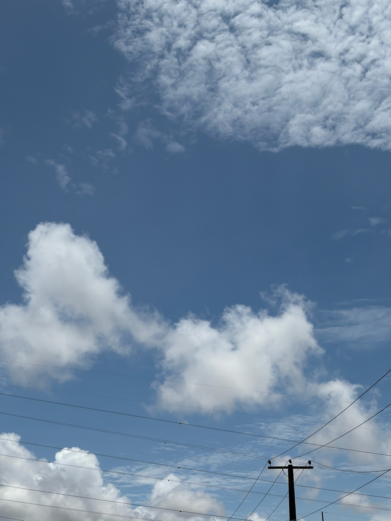 In Igboefon, Nigeria, a sky of clear blue is dotted with wispy white clouds. The lower portion of the image features several power lines stretching horizontally across the frame, with a wooden utility pole visible on the right, supporting some of these lines. The clouds are varied, with some appearing soft and scattered, while others are denser and more defined, adding depth to the sky. The overall scene captures a typical day in an urban or semi-urban environment, with the infrastructure of power lines juxtaposed against the natural backdrop of the sky and clouds.