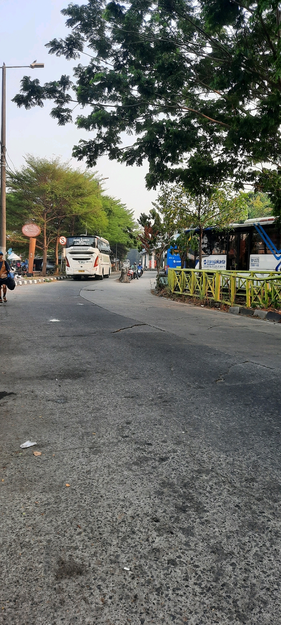 The image shows a street scene in East Jakarta, Indonesia, likely near a bus terminal or transportation hub. In the foreground, the asphalt road is visible with some cracks and debris. On the left side of the road, a person is walking with a dark bag. Further down the road, a white bus is parked, and a blue TransJakarta bus is visible on the right side of the road, with a yellow fence in front of it. Trees line the street, and there are buildings and signs in the background. Several motorcycles are also present on the road. The sky is overcast, suggesting it may be early morning or late afternoon.