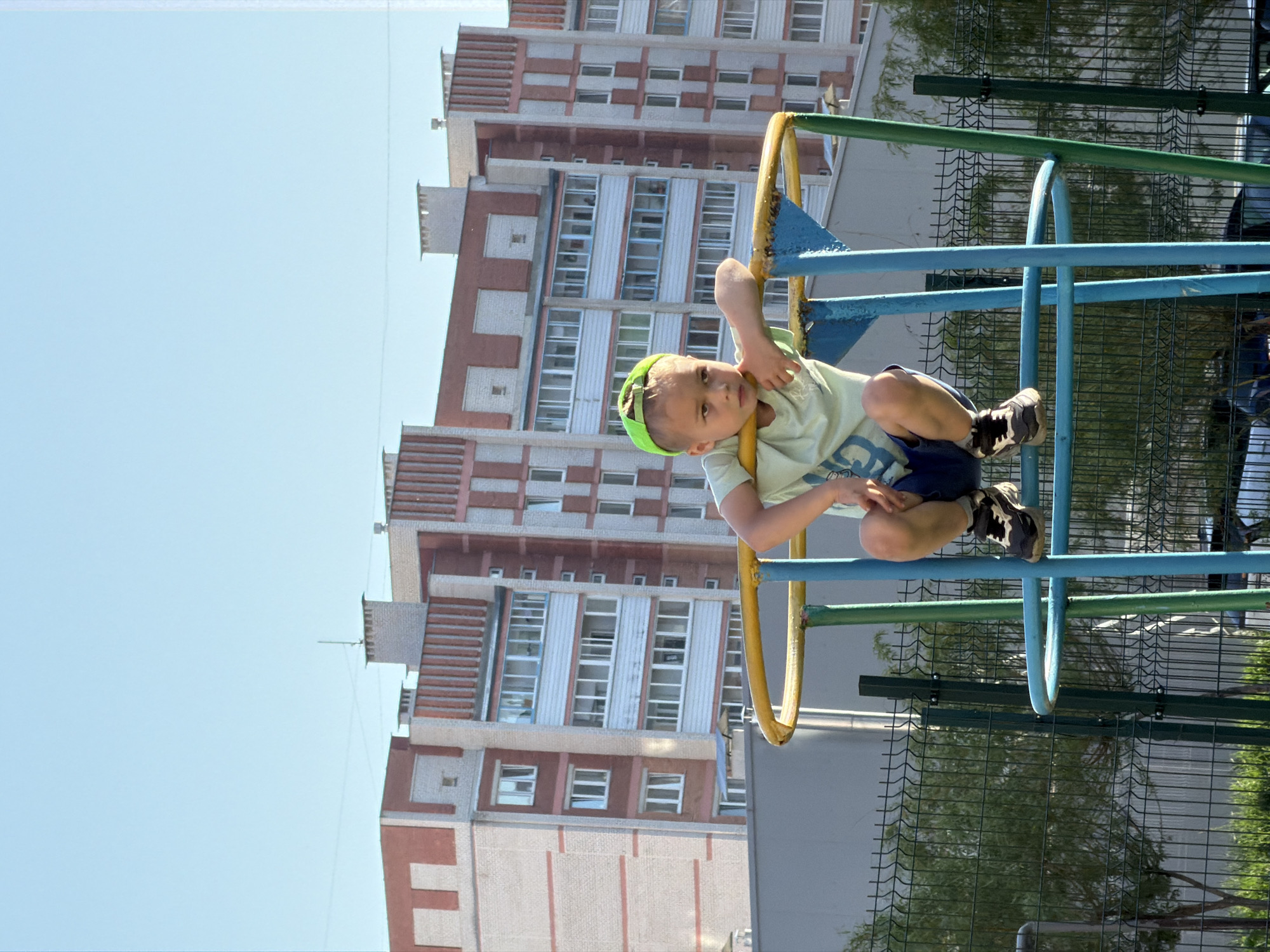 A child wearing a green cap, light green shirt, and dark shorts is sitting on a blue and yellow metal playground structure. The child is gripping the structure with their hands. Behind the playground, a tall building with white and red brick accents rises into the clear blue sky. A green fence and trees are visible in the background to the right of the playground. The photo appears to have been taken during the day in Unknown, Unknown.