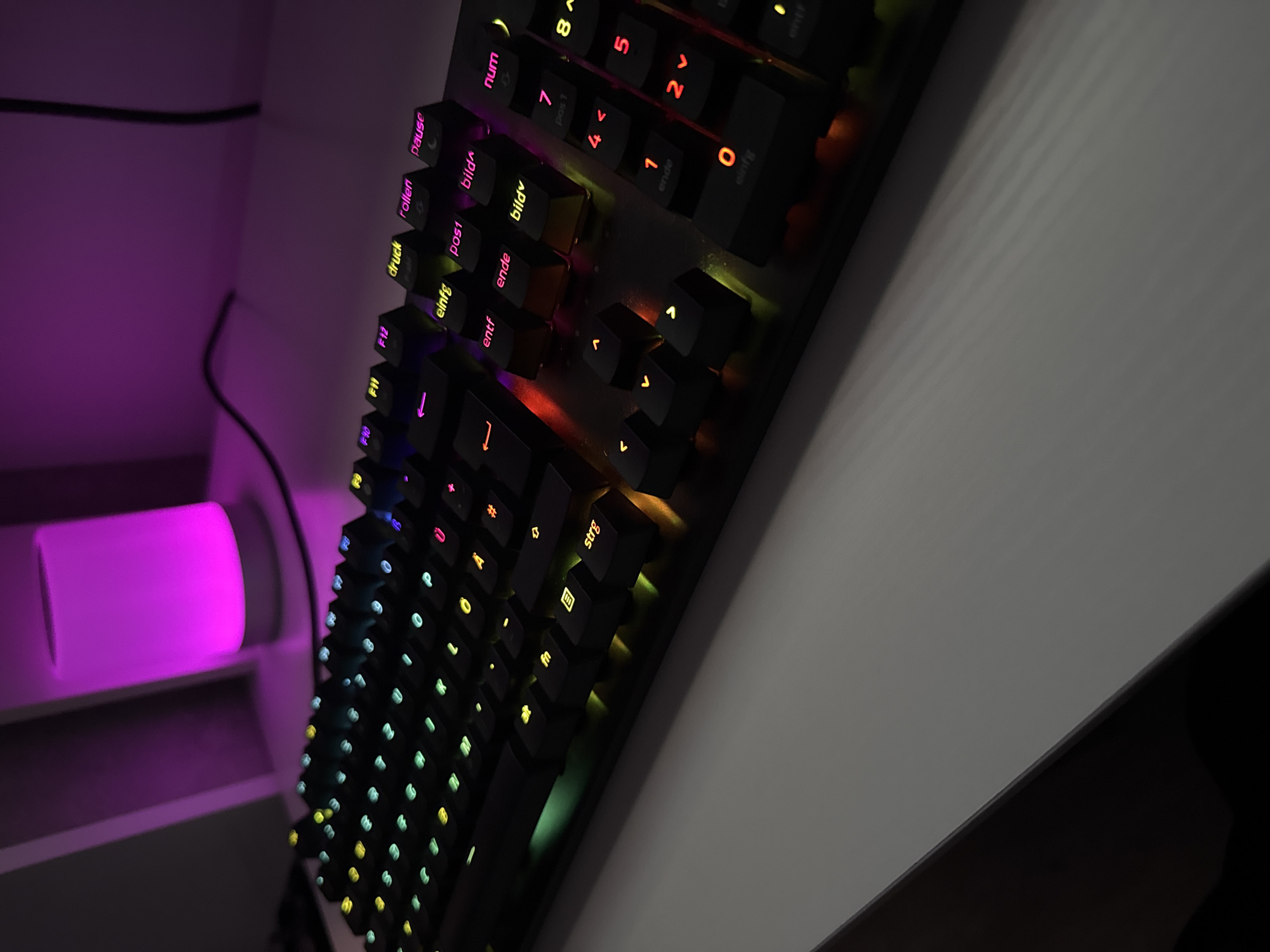 The image shows a close-up, high-angle view of a black mechanical keyboard with customizable RGB backlighting, illuminating the keys in various colors including green, yellow, pink, and orange. The keyboard is positioned on a white, textured surface, likely a desk. In the background, a cylindrical lamp emits a vibrant pinkish-purple glow. The scene is indoors, with a dark background suggesting a controlled lighting environment. There are no people visible in the image, nor any specific landmarks that would place it in Lübeck, Germany. The focus is on the illuminated keyboard and its aesthetic presentation.