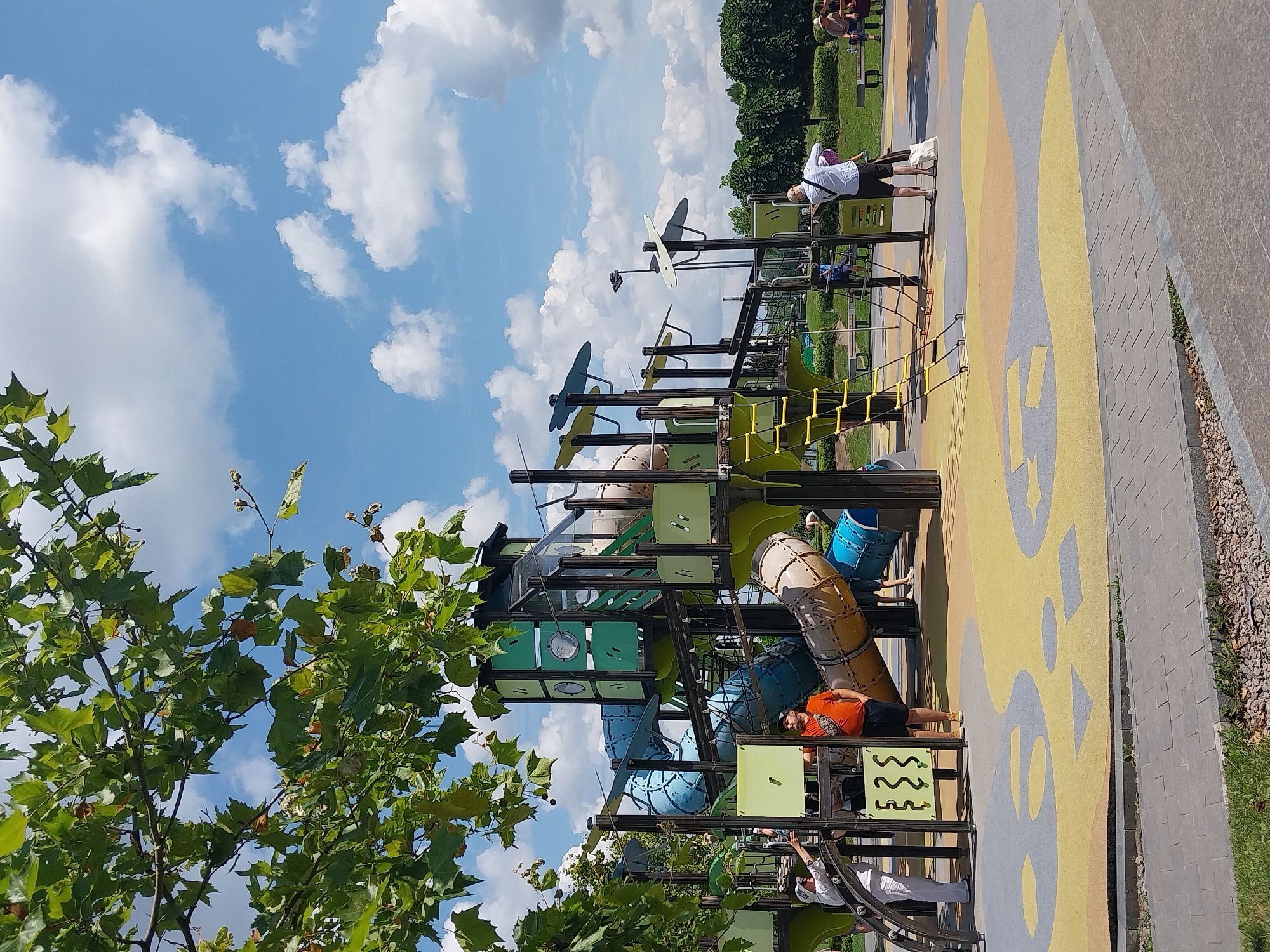 In Dnipro, Ukraine, a vibrant playground is depicted on a sunny day with a partially cloudy sky. The playground features a large, multi-level structure with blue, green, and tan tubular slides and climbing elements. A yellow ladder leads up to one of the platforms. The ground surface of the playground is a colorful, cushioned mat with abstract yellow, grey, and orange patterns.
Several people are present. In the foreground, a man in an orange t-shirt and black shorts stands near the playground structure, looking towards the camera. To his left, a person in a white outfit is partially visible, also near the structure. Further in the background, on the right side of the image, several individuals are seated on benches, with some children playing in the distance. Lush green trees and bushes surround the playground, and a paved walkway is visible in the foreground. The overall scene captures a lively atmosphere at a public recreational space.