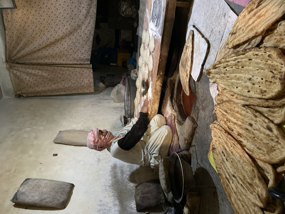 An individual, wearing light clothing and a head covering, is seated beside a wall-mounted oven. The person appears to be pressing dough circles onto the oven's surface. Stacked flatbreads lean against the wall nearby, indicating a batch of freshly baked goods. The setting is a simple room with a concrete floor. A patterned curtain partially obscures an adjacent area.