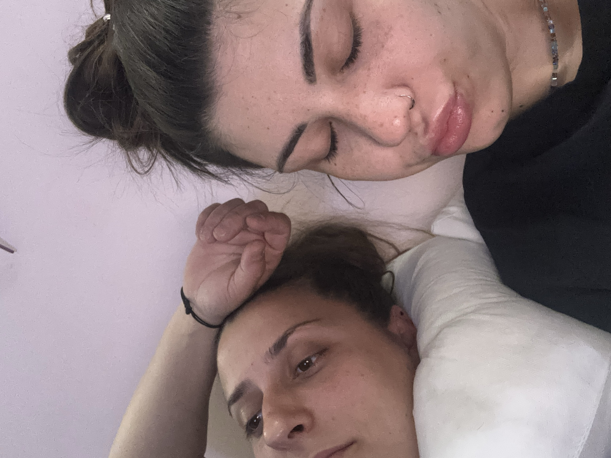 Two women are lying down. One woman's eyes are closed, and she has freckles, a nose ring, and a beaded necklace. Her hair is in a bun. The second woman is looking towards the camera, with her arm raised above her head. Her hair is also in a bun, and she wears a black bracelet. They are on a white pillow. The background is a light lavender color. The image was likely taken indoors, possibly in a bedroom, but there's no information to suggest the location is in the city of Sofia, Bulgaria.