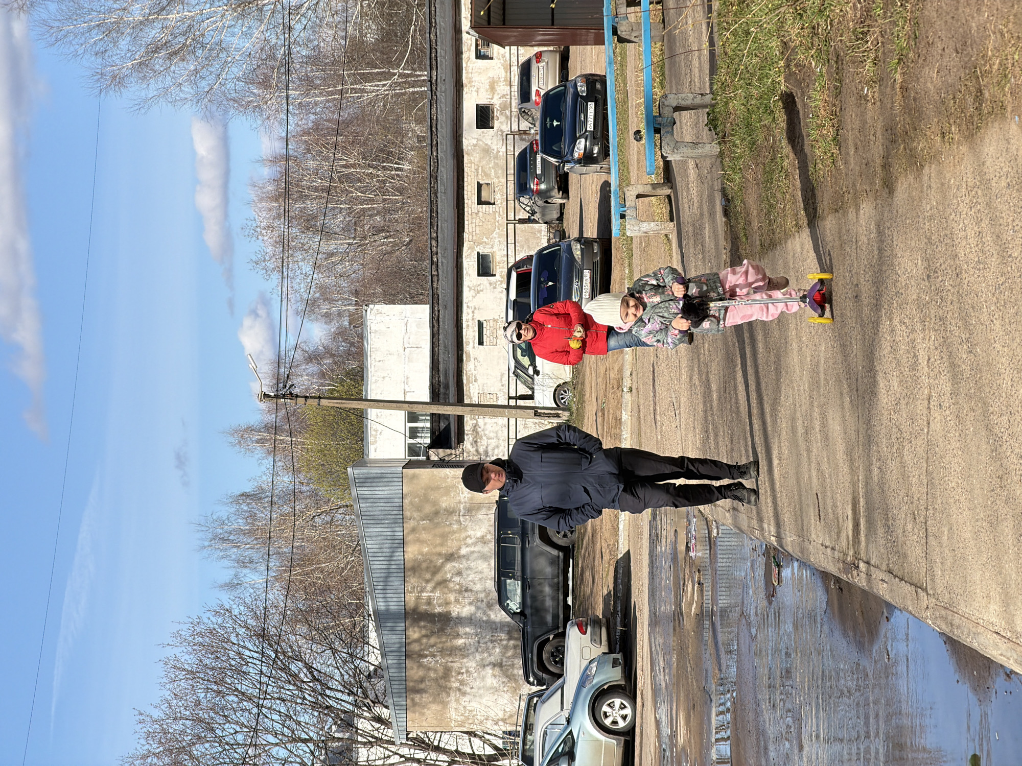 The image depicts a street scene in the city of Chaikovsky, Russia. On the right side of the street is a sidewalk where a man in a dark blue jacket and black pants is standing. Further down the sidewalk, an adult in a red jacket and sunglasses is standing next to a child on a scooter. Behind them is a building with parked cars. On the left side of the image, there is a puddle reflecting the sky and the surrounding buildings. There are more parked cars and bare trees visible in the background. The sky is blue with some clouds. Overhead power lines are strung between poles. The scene appears to be taken during the day in early spring.