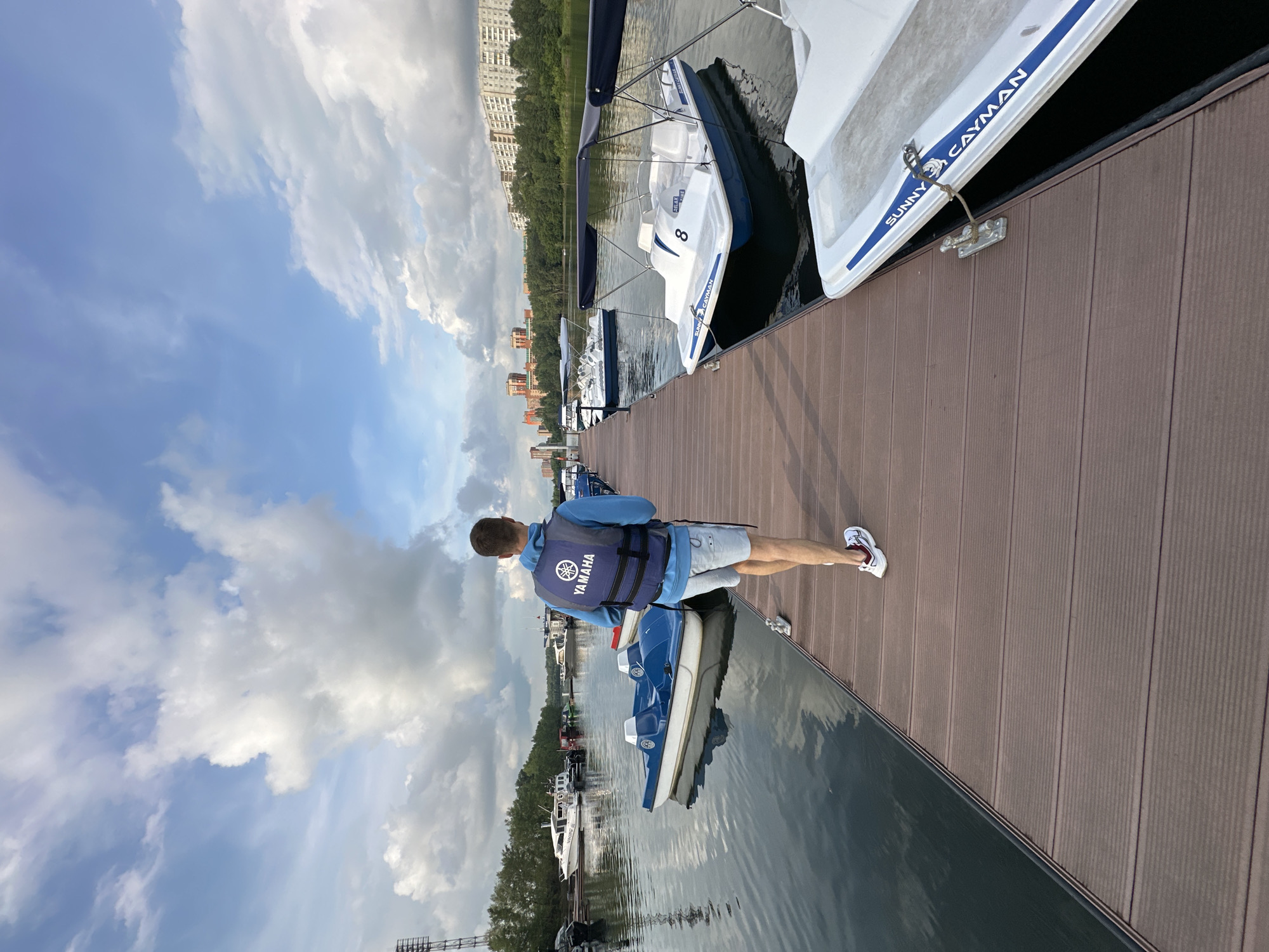 A man wearing a blue hoodie and a Yamaha life vest walks away from the camera on a wooden pier. He has on light-colored shorts and white athletic shoes with red accents. The pier is lined with several blue and white pedal boats with canopies. In the background, there are more boats docked, as well as buildings and trees. The sky is blue with scattered white clouds.