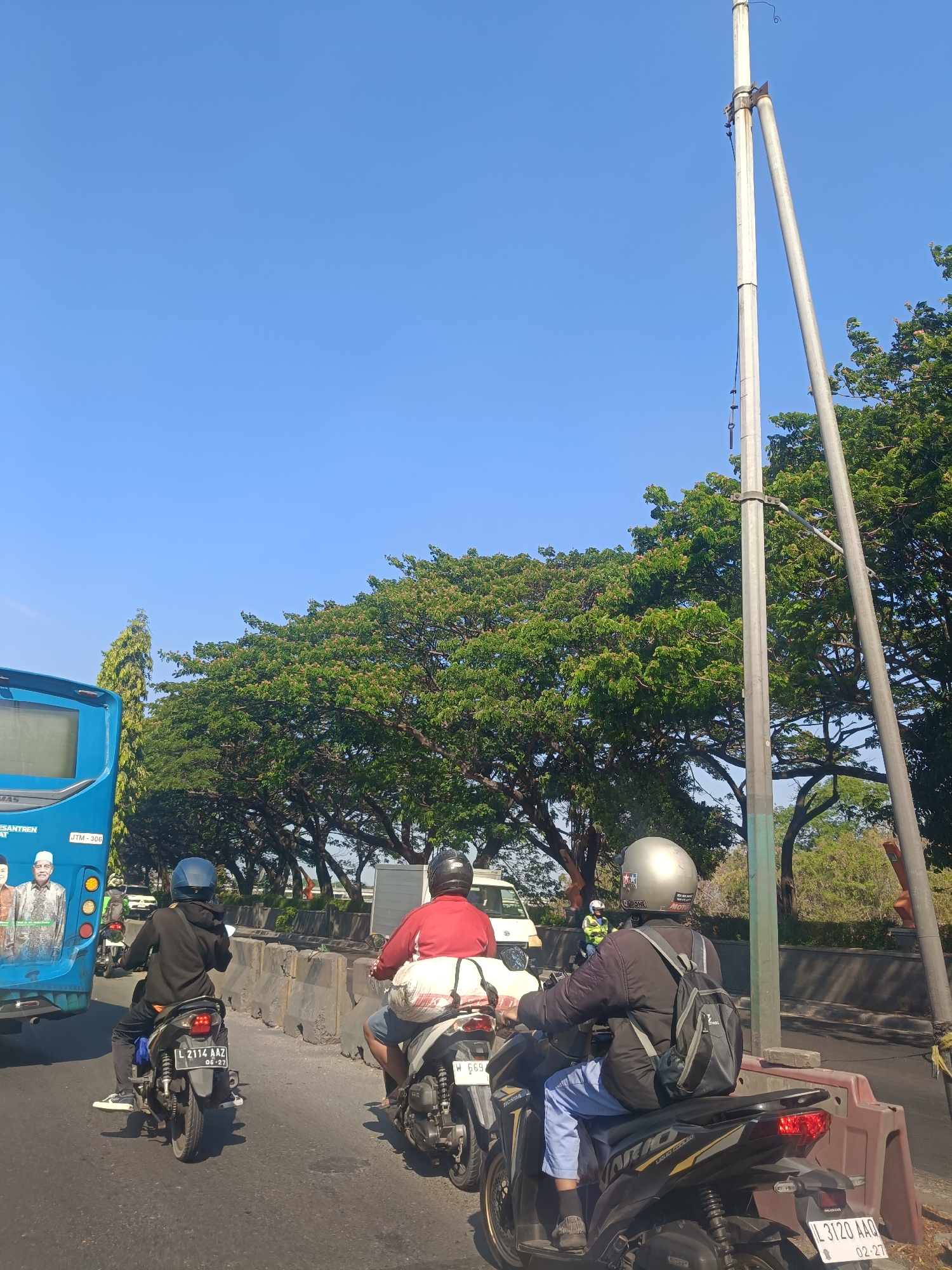 In the city of Dahanrejo, Indonesia, on a clear day with a bright blue sky, a traffic scene unfolds. A blue bus with images of men on its side is parked on the left. Several motorcycles are on the road. One motorcycle, black and silver, is in the foreground, with a person wearing a black jacket, blue pants, and a silver helmet riding it. Another motorcycle, white, is next to it, ridden by a person in a red shirt and blue shorts, carrying a white bag. The road is lined with concrete barriers and trees. There are utility poles on the right side of the image.