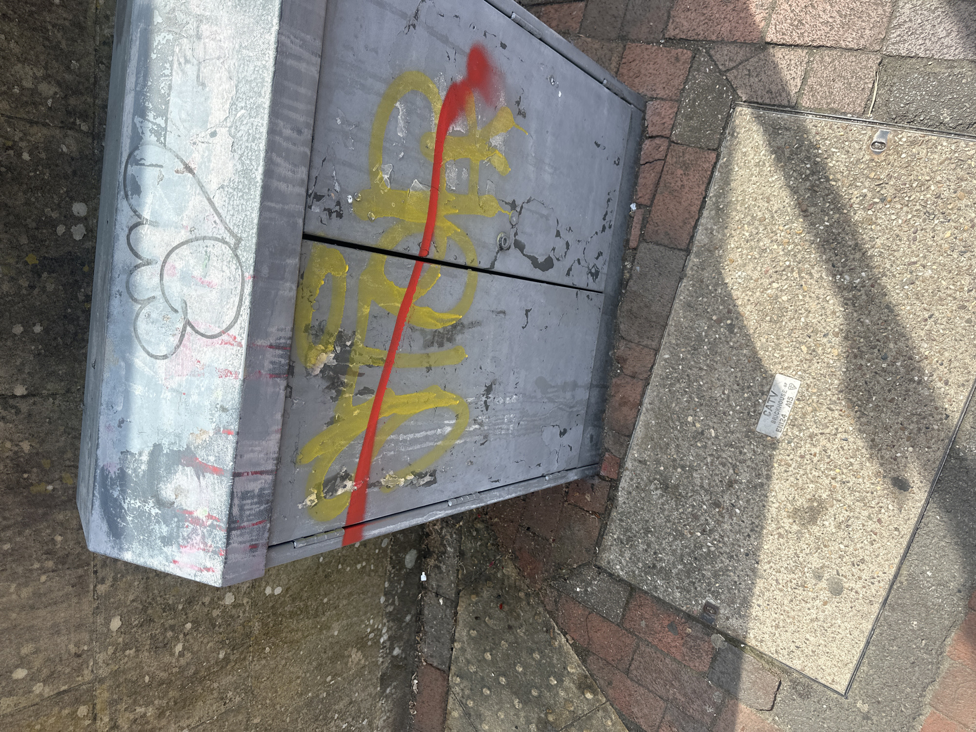 An outdoor image shows a gray metal utility box with graffiti in yellow and red paint. The graffiti appears as abstract, overlapping shapes. To the right of the box is a concrete square utility access cover with a label indicating "CATV BRICKHOUSE," potentially Tunbridge Wells, UK. The surrounding ground consists of various textures, including concrete, asphalt, and brick. The lighting is bright and casts shadows on the ground.
