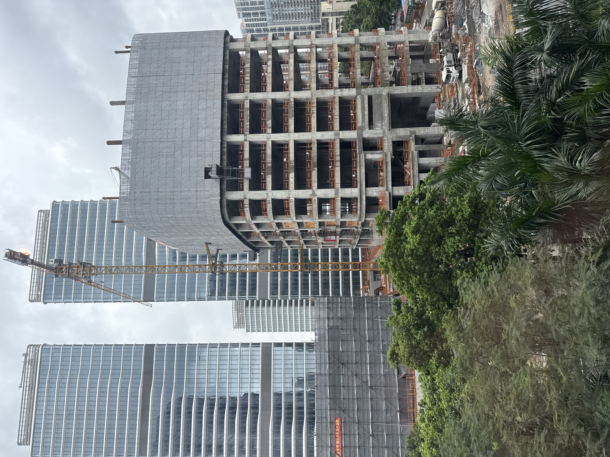 The image depicts an urban construction site in Haizhu District, China, under a cloudy sky. Two completed modern skyscrapers with glass facades stand to the left and in the background. In the foreground, a multi-story concrete building is under construction, partially covered by a large gray mesh. A yellow construction crane is positioned between the skyscrapers, extending over the unfinished building. The ground level of the construction site shows piles of debris, building materials, a cement truck, and a white truck. Lush green trees, including palm trees, are visible in the lower portion of the image, suggesting a tropical or subtropical environment. Some scaffolding is also present, along with construction barriers and signage. No people are clearly visible in the image.