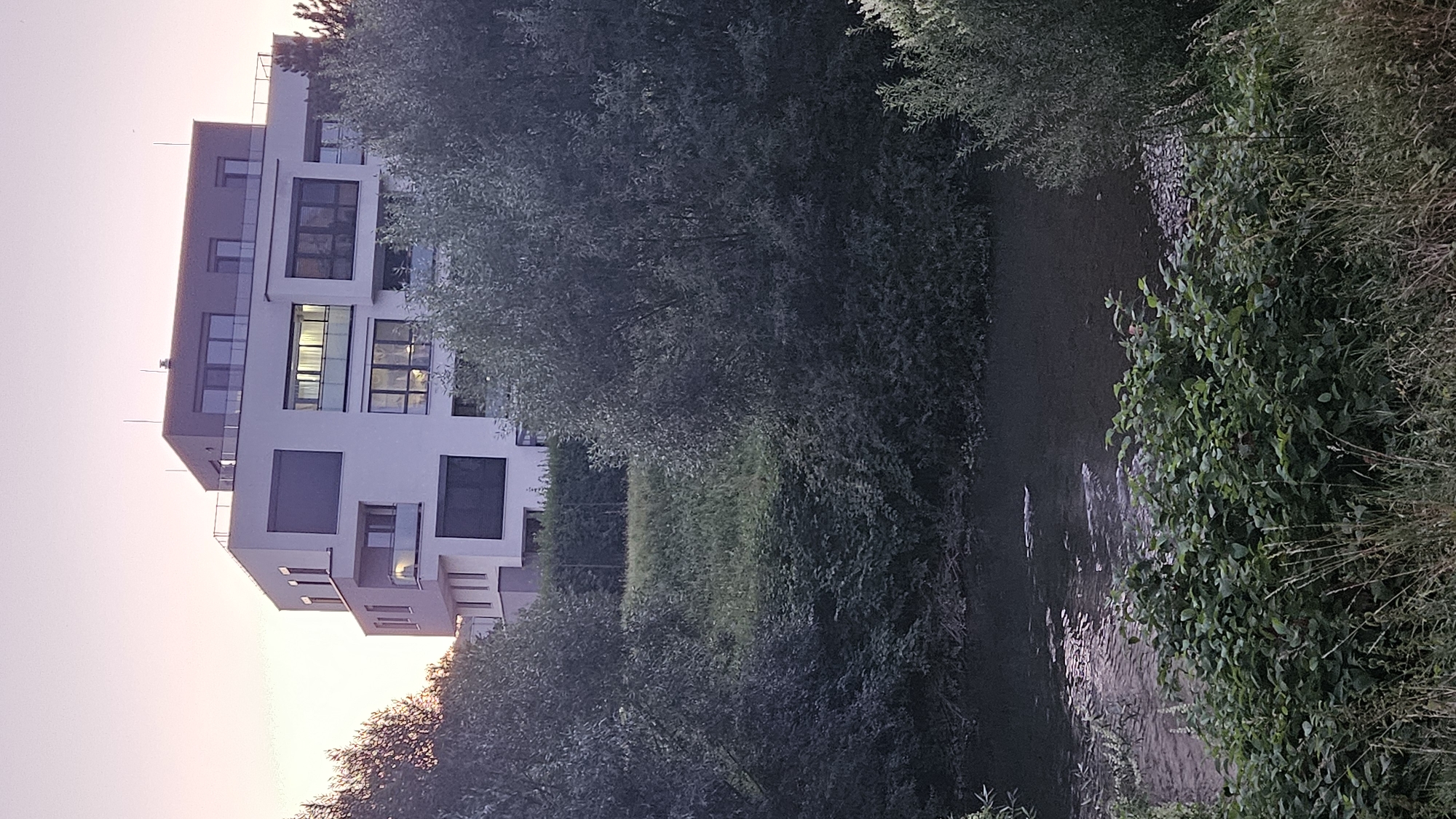 The image captures a modern white apartment building with dark-tinted windows and balconies, situated on a slight incline. The building has multiple levels and a flat, dark-colored roof with glass railings. In front of the building, there is dense greenery, including trees and bushes, and a body of water, possibly a river or stream, flows in the foreground. The sky is a soft, pale pink, suggesting it is either dawn or dusk. The image is taken from a perspective at ground level, looking upwards towards the building and across the water. The scene appears to be a residential area, likely on the outskirts of a city, with a blend of urban architecture and natural landscape. The location is identified as Prešov, Slovakia.