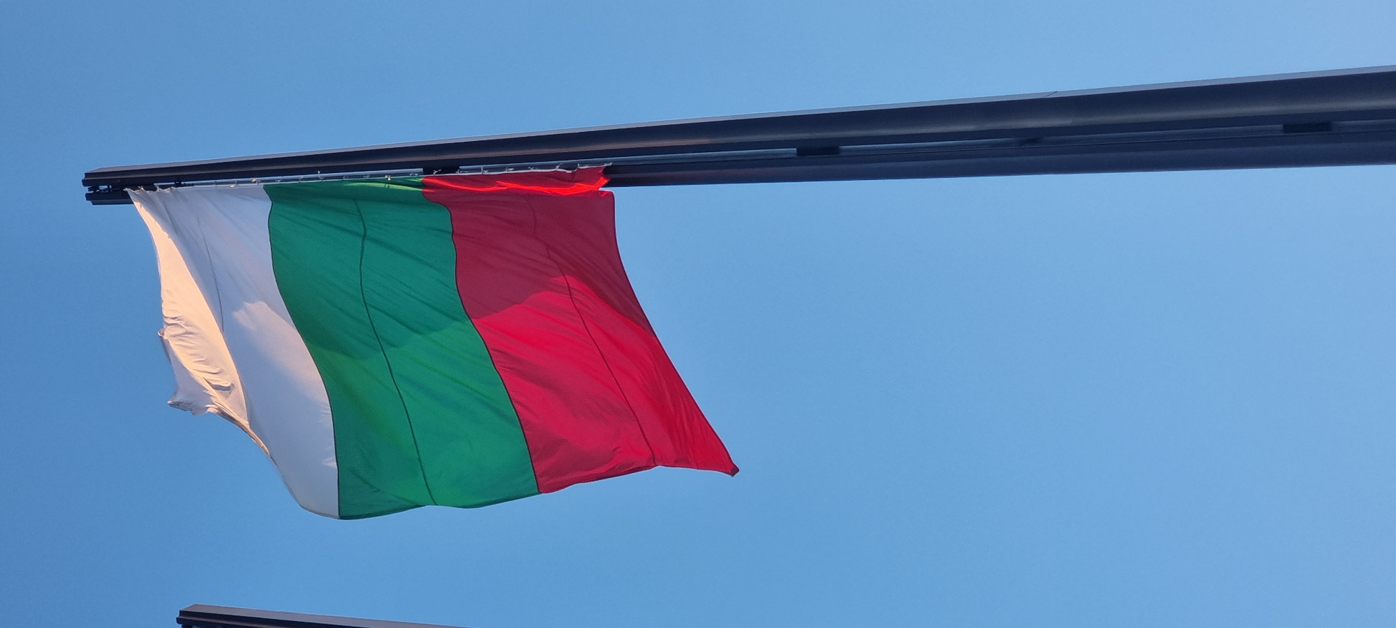 The image shows a Bulgarian flag waving against a clear blue sky. The flag is attached to a horizontal metal bar. The three stripes of the flag, white, green, and red, are clearly visible. The flag is slightly wrinkled, suggesting it is blowing in the wind. The scene is in Sofia, Bulgaria.