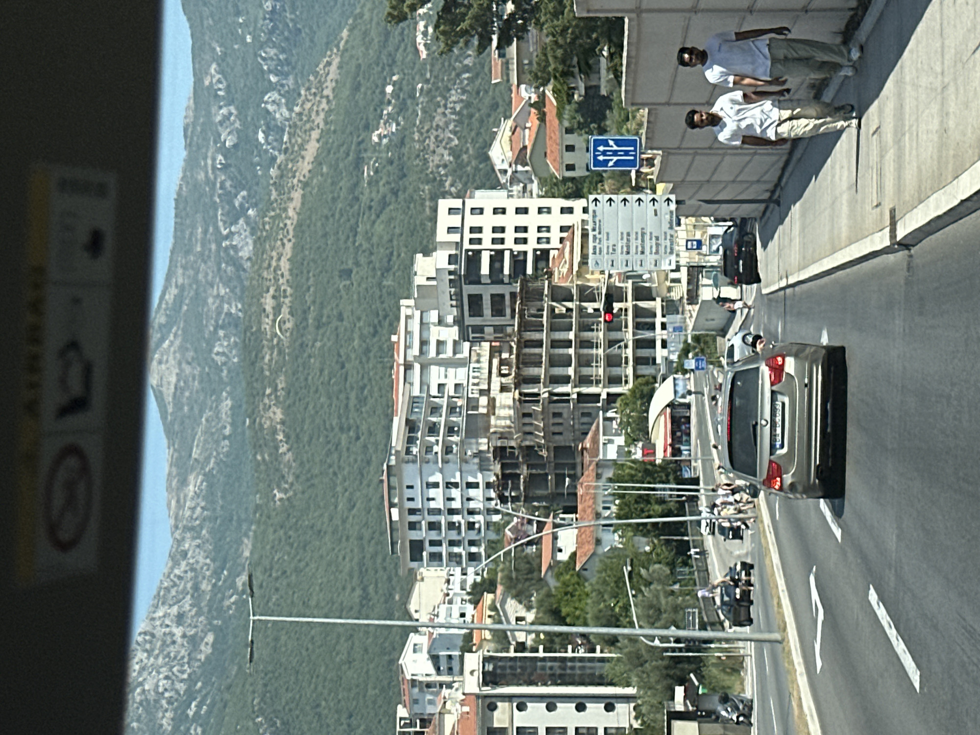 The image captures a street scene in Boreti, Montenegro, with a focus on a multi-lane road leading through a built-up area. In the foreground, a silver station wagon is driving down the right lane of the road. The road is marked with white lane lines, and an arrow indicates the direction of travel. To the right of the road, a sidewalk runs parallel, and two men are walking in the same direction as the car. They are both wearing casual attire: one in a white polo shirt and beige pants, the other in a light-colored t-shirt and beige pants.
Beyond the sidewalk, there is a light-colored concrete wall. Further back, several multi-story buildings are visible, including a large white building with many windows and a building under construction with exposed concrete and scaffolding. A street light is prominently positioned on the left side of the road. In the background, lush green hills rise, with a clear blue sky overhead. Some scattered residential buildings can be seen on the hillsides. A paraglider is visible as a small speck in the sky above the hills. Traffic signs are posted on poles near the buildings, indicating directions to various locations. The overall impression is of a sunny day in a coastal town with a mix of modern architecture and natural landscapes.