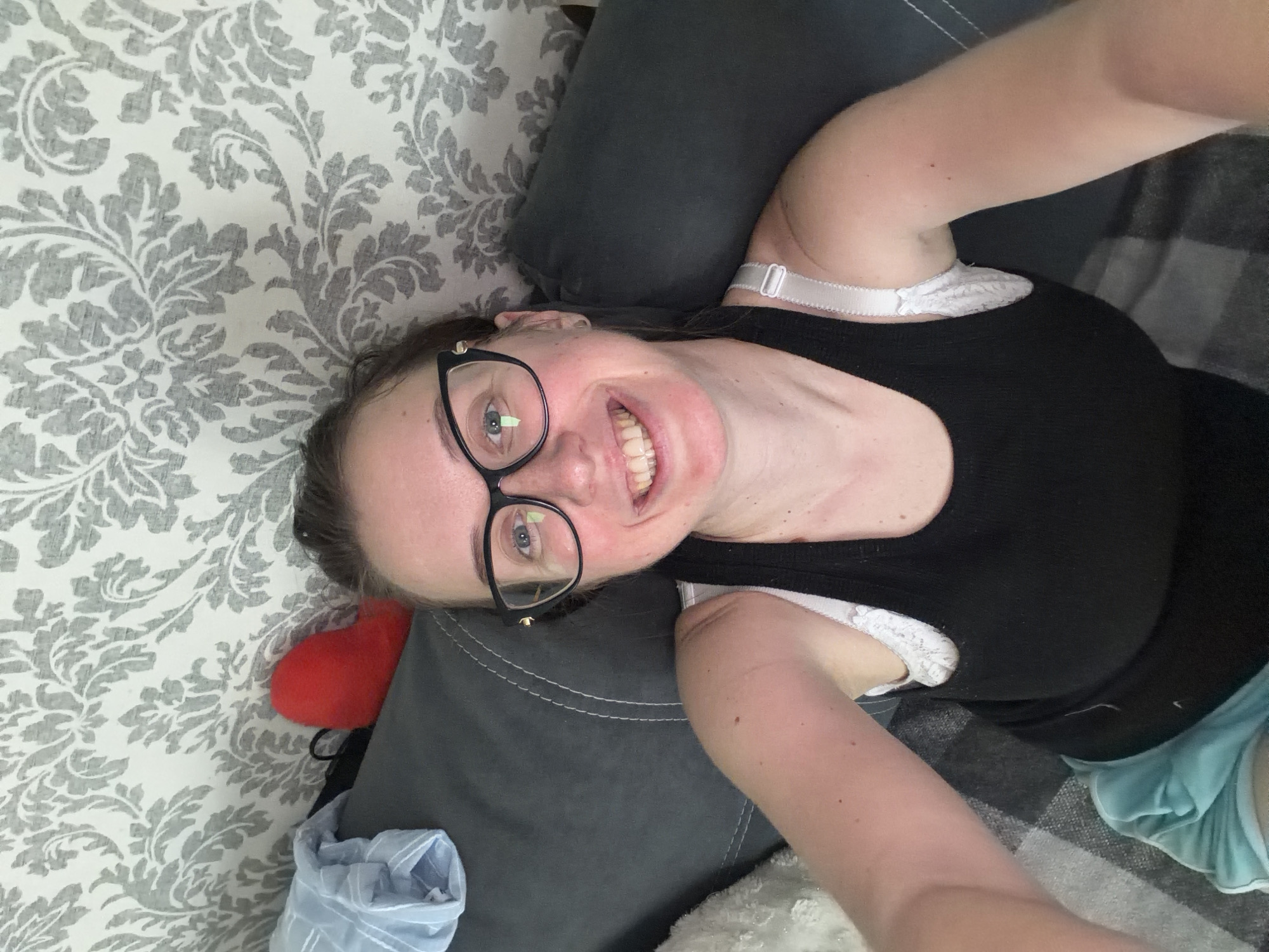 A medium shot captures a young woman with fair skin, blonde hair pulled back, and glasses with black frames, smiling as she looks directly into the camera. Her eyes are blue, and her teeth are visible in her smile. She is wearing a black tank top and a white lace bra with visible straps. The background shows a grey sofa with white stitching and a patterned wallpaper in shades of grey and white. To the left of the sofa, a folded light blue garment and a small red object are visible. The lighting suggests an indoor setting, possibly a living room. The woman appears relaxed, lying on the sofa. The provided crop images confirm the details of the woman's face, upper body, and the surrounding environment. There is no information present in the image or crops to indicate a location in Bila Tserkva, Ukraine.