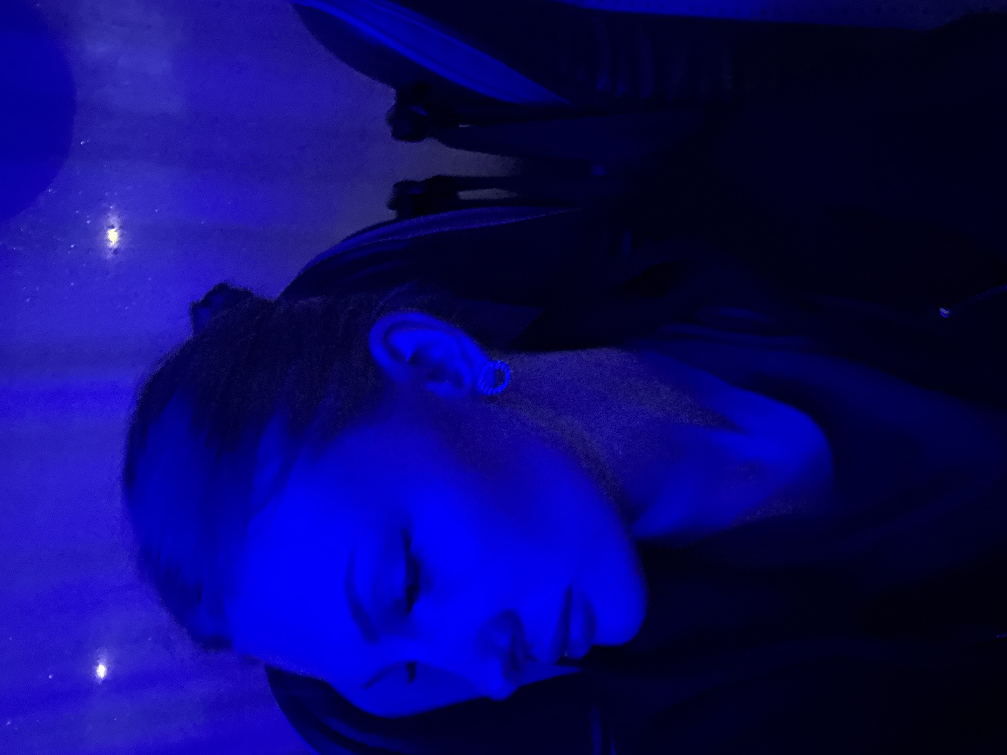 A portrait of a young woman is captured in a dimly lit room with predominantly blue lighting. The woman is shown from the side profile, her head tilted down and her eyes closed. She has her hair pulled back in a bun. A small, circular earring is visible in her left ear. The background appears to be the interior of a theater or similar venue, with seats visible behind her. The overall mood is contemplative and serene. The image does not contain any specific landmarks or indicators that would allow for the identification of the city of Kazan, Russia.