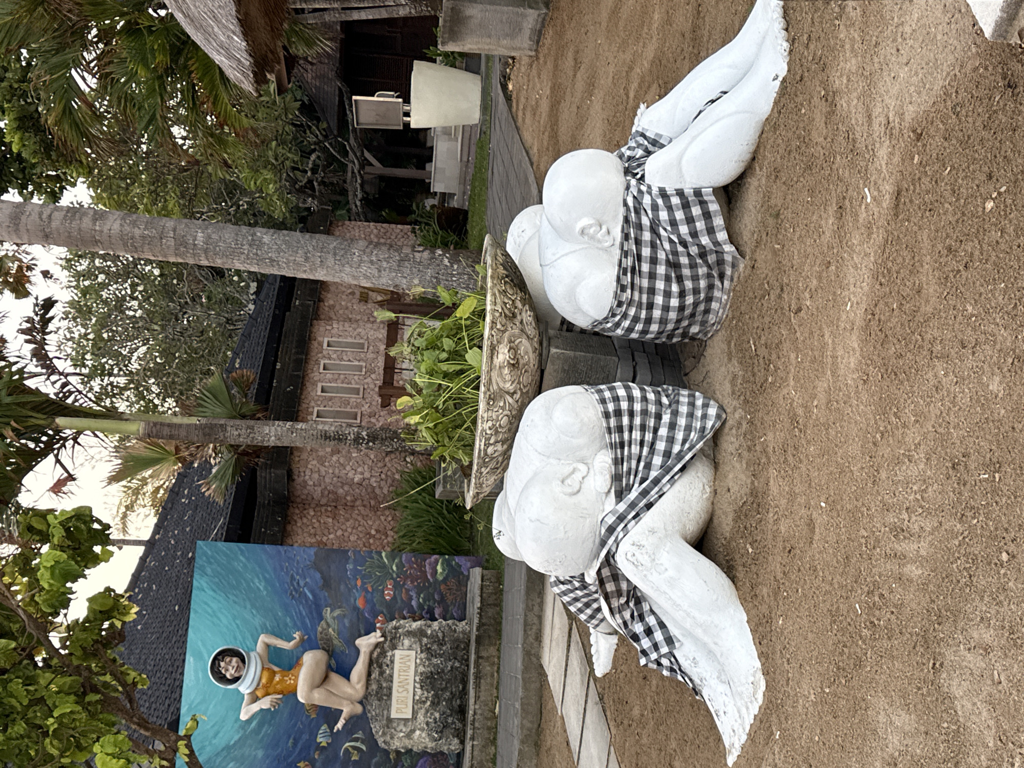 The image depicts an outdoor scene at Puri Santrian, a resort in Denpasar, Indonesia. In the foreground, two white, bald, reclining statues are draped with black and white checkered fabric. They appear to be resting or in a somber pose, with their heads bowed. Behind the statues, a large planter filled with green foliage sits atop a stone base. Further back, a colorful mural on a wall features a woman in a diving suit in an underwater scene with fish and turtles. The mural is labeled "PURI SANTRIAN". The environment includes sandy ground, paving stones, trees, and buildings with thatched roofs, suggesting a tropical resort setting. The lighting indicates it is either early morning or late afternoon.