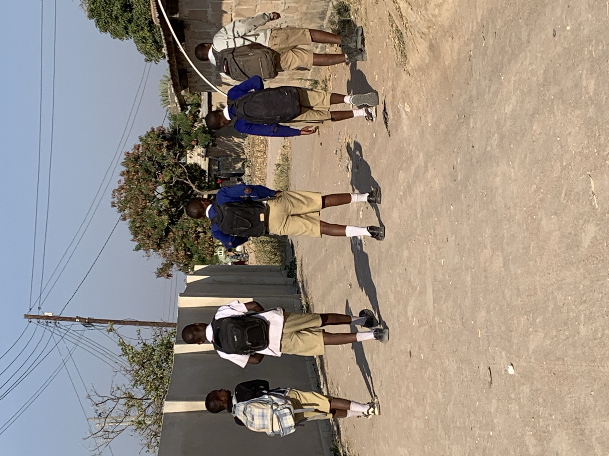 The image depicts five young males walking away from the camera on a dirt road in Dodoma, Tanzania. They are all wearing school uniforms consisting of light-colored shorts and shirts or long-sleeved shirts. Each of them is carrying a backpack. The environment is characterized by a dirt road, a low concrete wall on the left, and buildings and vegetation in the background. Power lines are visible overhead, suggesting an urban or semi-urban setting. The lighting indicates it is daytime, likely late morning or early afternoon, with clear skies.