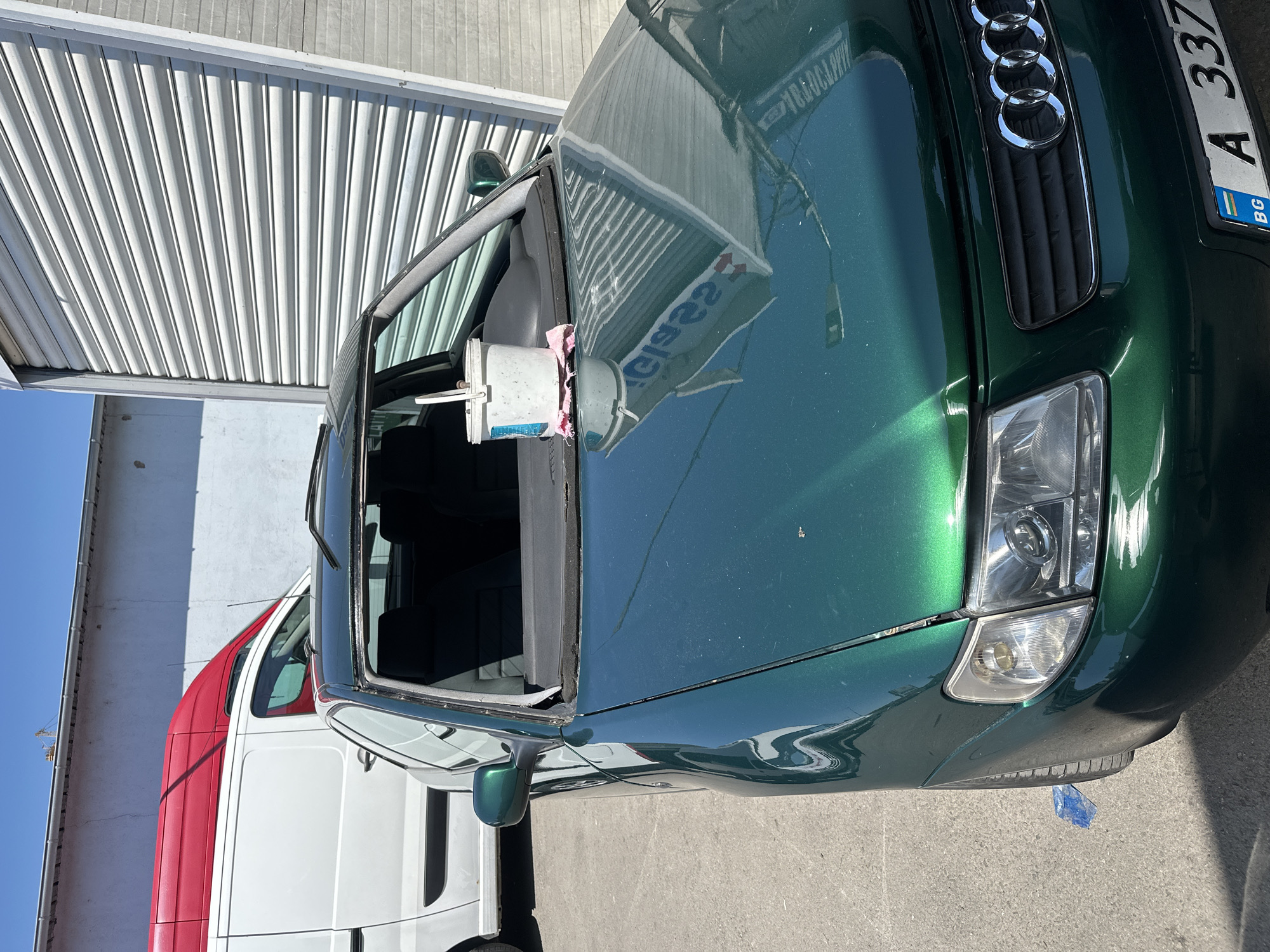 A dark green Audi A3 is parked on an asphalt surface in front of a large, ribbed metal garage door. The car's windshield is missing, and a white bucket with a pink cloth is placed on the dashboard. A white van with a red roof is parked to the left of the Audi. The sky is clear and blue.