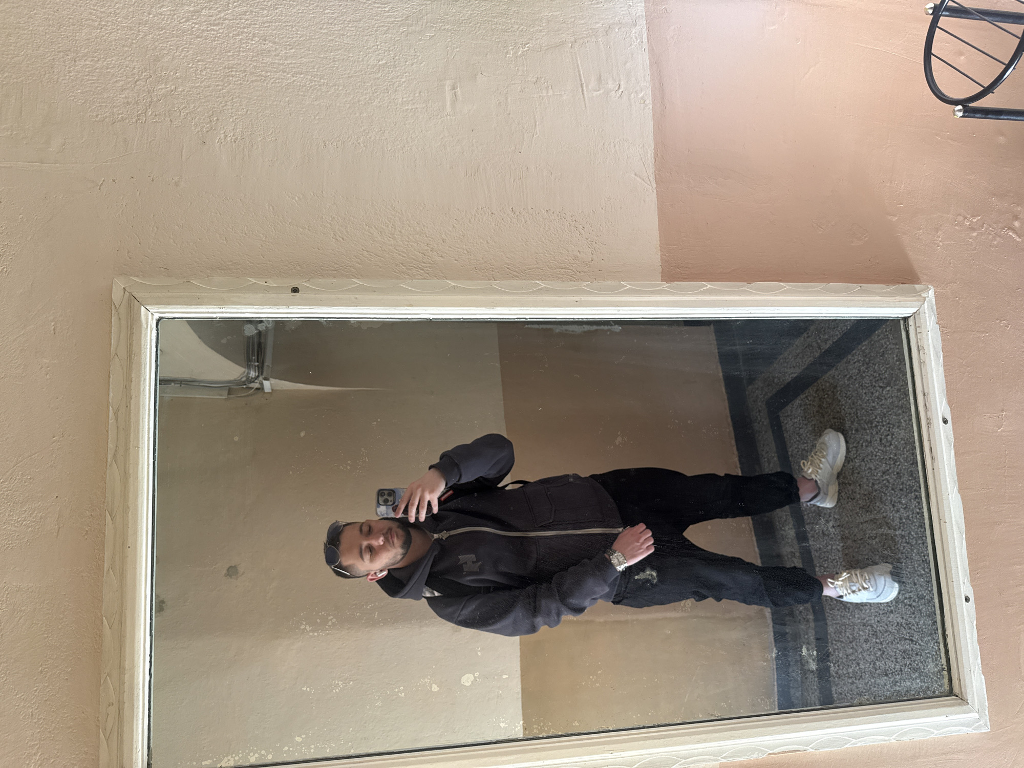 A person is taking a selfie in a rectangular mirror with a decorative white frame. The mirror is mounted on a wall with a two-toned paint job. The wall is pale yellow-beige on the left and pale rose-beige on the right. The person is wearing a dark hooded jacket and dark pants, with white sneakers. The reflection shows a tiled floor and a partial view of a pipe near the top left corner of the mirror. The location is likely indoors, but there are no clear indications of a specific building or location within Sofia, Bulgaria.