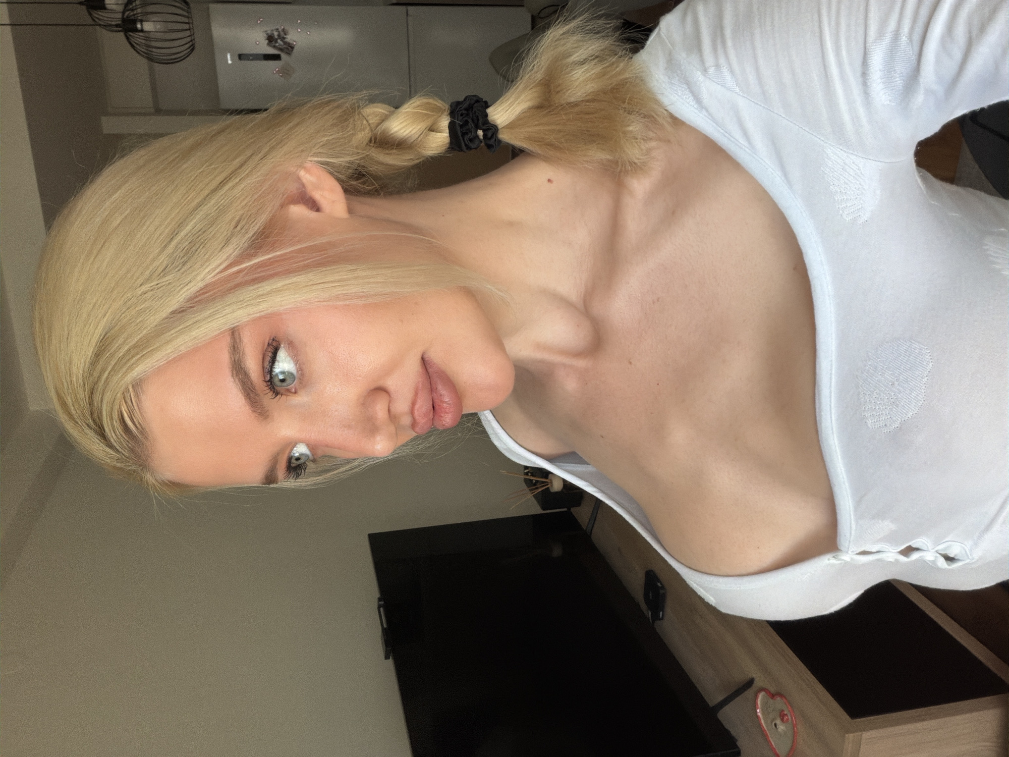 A young Caucasian woman with blonde hair and light-colored eyes is depicted indoors, likely posing for a photograph. Her blonde hair is styled in two pigtail braids, secured with black scrunchies, with some strands framing her face. She has subtle makeup, including defined eyelashes. She is wearing a white, long-sleeved top featuring a subtle, textured pattern of circles or dots. Her head is tilted, and she looks towards the camera with a neutral to slightly soft expression.
The setting appears to be a room, possibly a bedroom or living area. In the background, a light-colored wall is visible, along with parts of dark furniture, which might be a television screen or a dark tabletop. Beneath it, a wooden-toned cabinet or shelf is present, on which a small red and white heart-shaped decorative object can be seen. Further in the background, an ornate, cage-like light fixture hangs from the ceiling. The lighting suggests it is daytime. There is no visible text in the image. The image provides no specific visual cues to indicate the location as Belgrade, Serbia, other than the provided context.