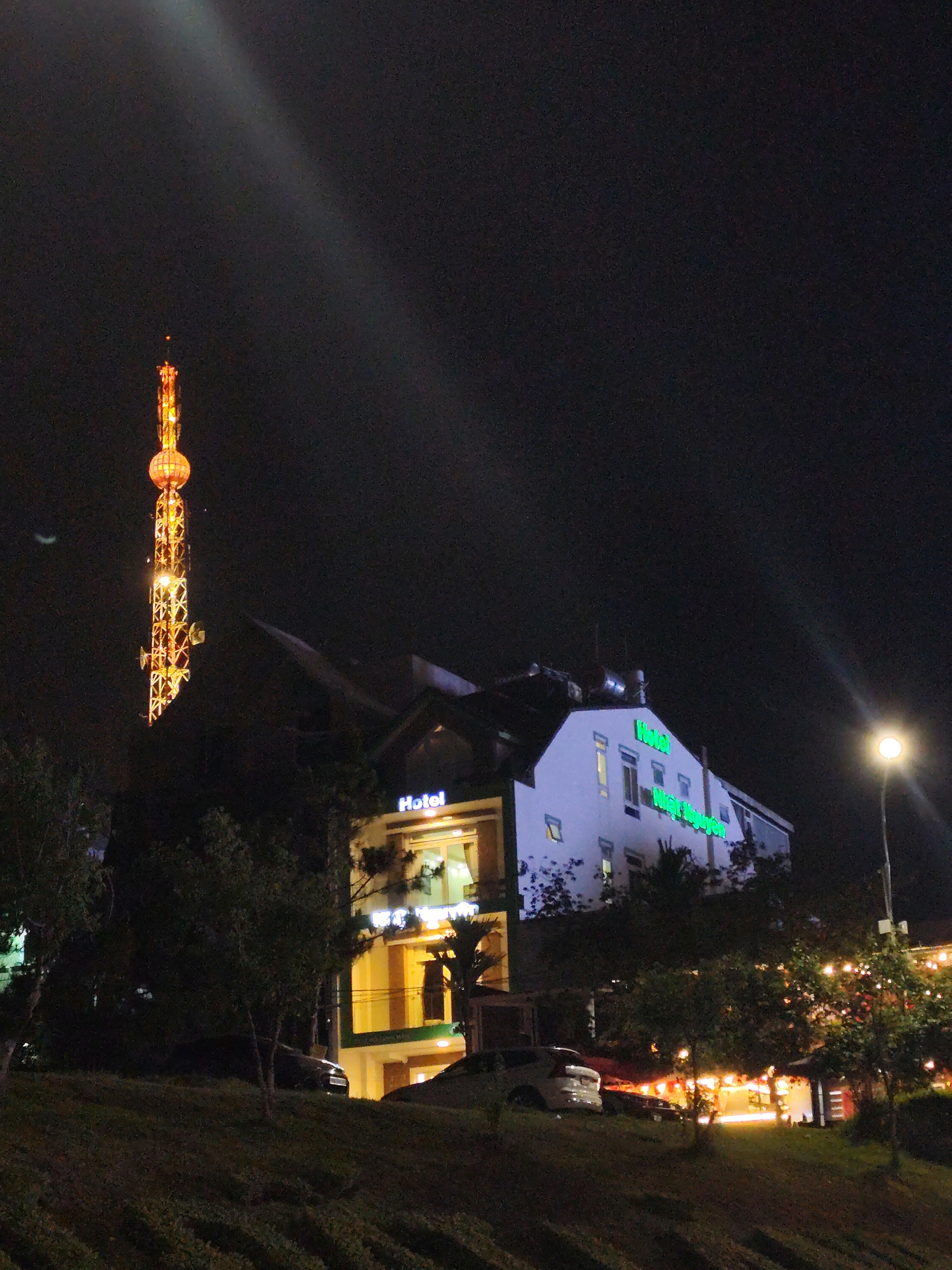 At night, a lit telecommunications tower stands to the left of a Hotel named "Hotel Nhat Nguyen" in Da Lat, Vietnam. The hotel is illuminated, as is a street lamp on the right. Cars are parked in front of the hotel on a sloping hill. Trees are scattered throughout the scene.
