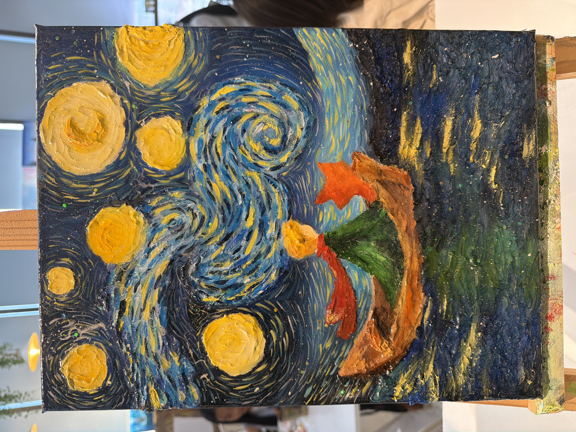 This is a painting of a scene inspired by Van Gogh's "Starry Night," featuring a dark blue night sky filled with swirling patterns of yellow and blue paint, representing the sky and stars. Several large, impasto yellow circles depict stars or celestial bodies. In the foreground, a figure is depicted, appearing to be a person in a green cloak with a red scarf, possibly the Little Prince from Antoine de Saint-Exupéry's novel. The figure is rendered in thick, textured paint, with an orange, curved shape behind them, possibly a planet or a stylized landscape element. The right side of the painting shows a dark, textured area with streaks of yellow, suggesting distant lights or a more abstract depiction of the night. The overall style is expressive and textured, with visible brushstrokes and the application of paint in thick layers. The painting is displayed on an easel, suggesting it is a work in progress or recently completed artwork. There is no visible text in the image. The context of Nha Trang, Vietnam, does not provide specific visual cues within the painting itself, as the imagery is a stylized interpretation of a celestial landscape. The emotion conveyed is one of wonder and perhaps contemplation, characteristic of the "Starry Night" theme and the Little Prince narrative.