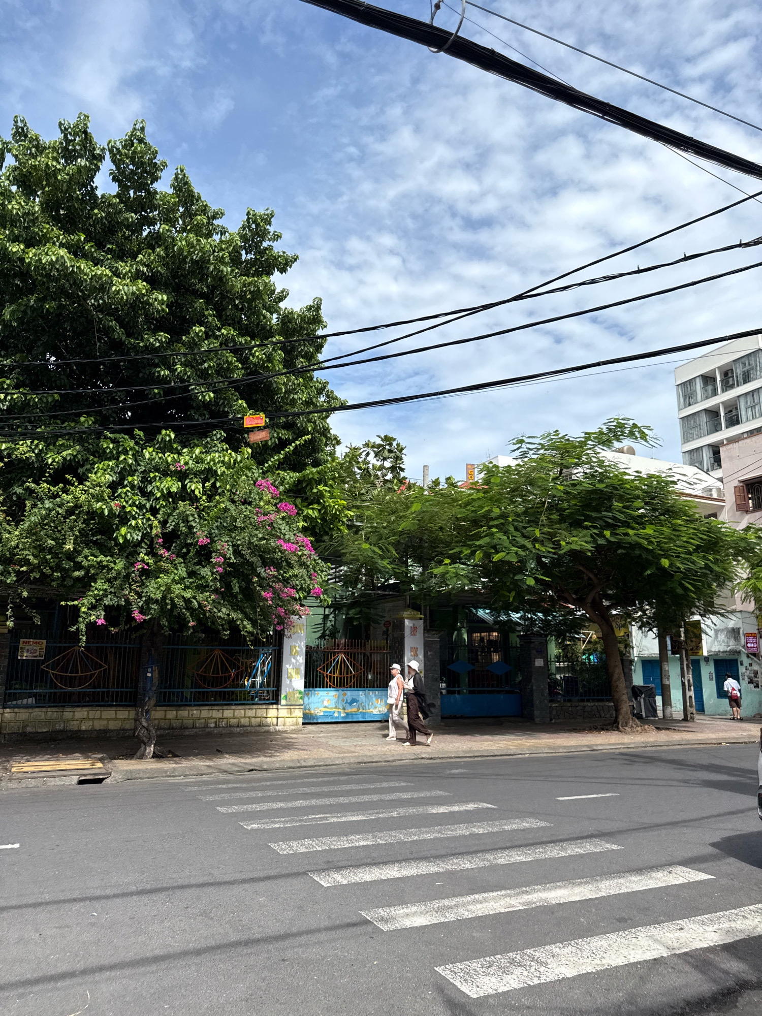 This image depicts a street scene in Nha Trang, Vietnam, featuring a pedestrian crossing in the foreground and a tree-lined sidewalk with buildings in the background. The sky is partly cloudy.
On the sidewalk, two individuals are walking from left to right. The person on the left is wearing a white hat and a light-colored top. The person on the right is wearing a dark hat and a dark outfit. Behind them, a large tree with green foliage and some pink flowers dominates the left side of the frame. A fence separates the sidewalk from a building with a blue facade and colorful decorations. To the right of this building, there are more trees and structures, including a multi-story building with windows. Utility wires are visible crisscrossing the upper portion of the image. The street itself is paved asphalt with white stripes marking the pedestrian crossing.