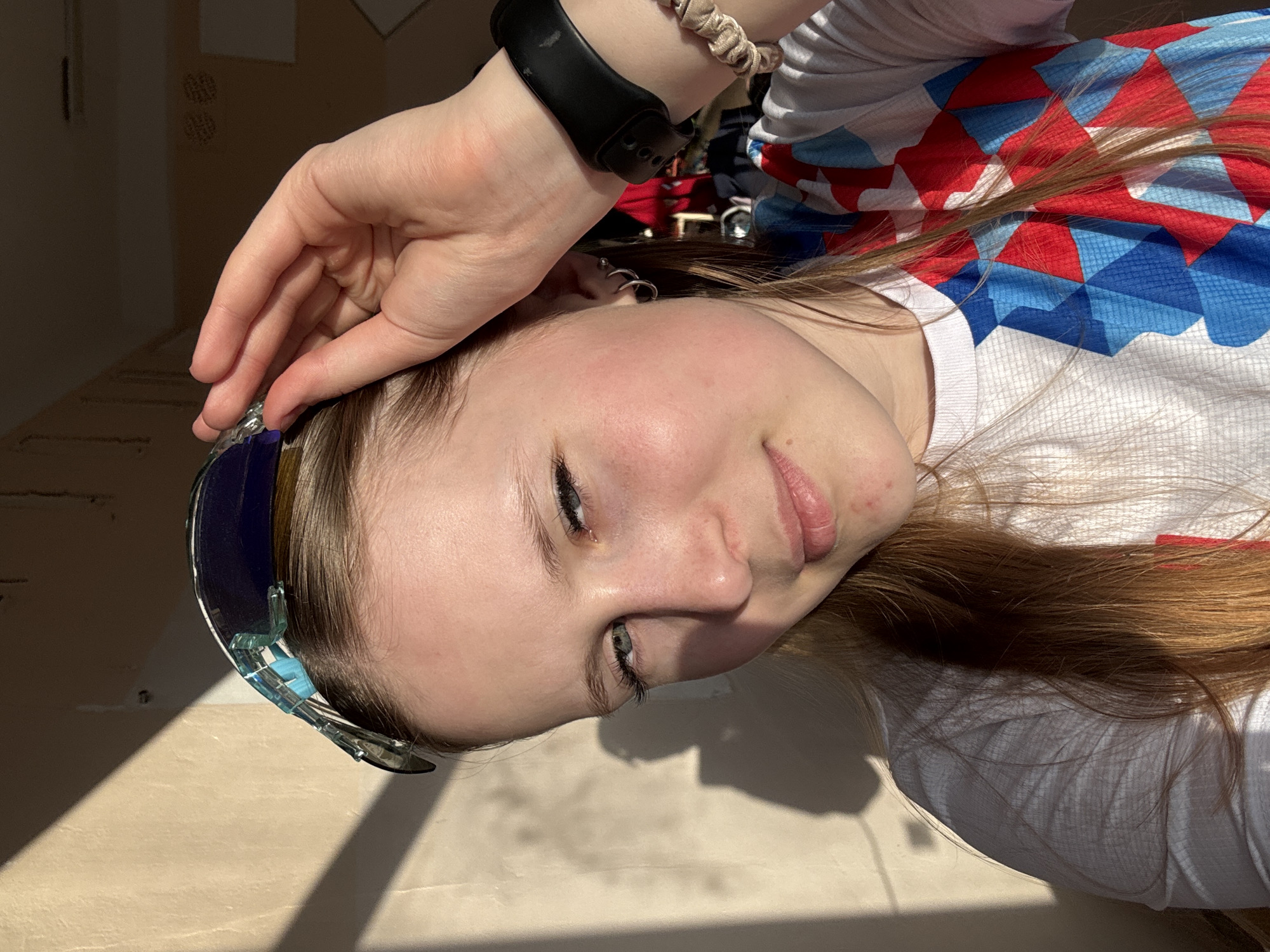 A young person with light skin, light brown hair, and blue eyes is lying down. The person is wearing a white long-sleeve shirt with red and blue geometric patterns. The person is wearing a black smartwatch on their left wrist, along with a tan scrunchie. The person is holding a pair of blue sports sunglasses. The location is unknown; there is no indication that the location is in Usolye, Russia.