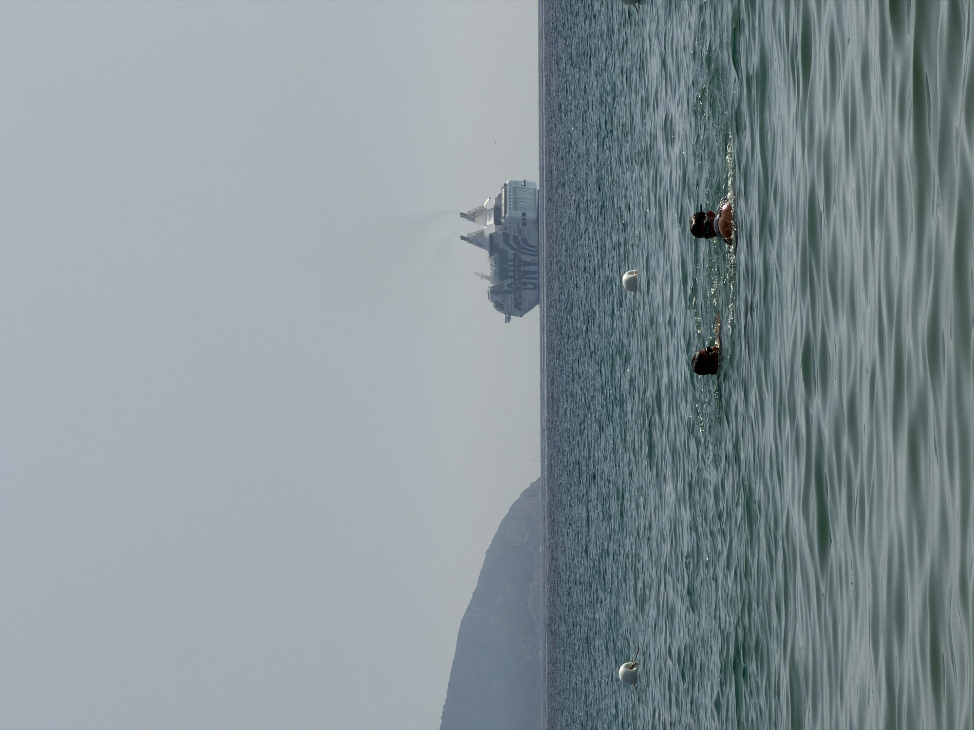 A wide-angle, outdoor, eye-level shot captures a serene seascape with a distant ferry and two swimmers in the foreground. The water is a deep green, with gentle waves creating a rippled texture. Two white buoys are visible, one in the left mid-ground and another in the right mid-ground. In the background, a large, light-blue ferry with dark blue lettering and white accents is visible on the horizon, emitting a plume of smoke from its two funnels. To the left of the ferry, a hazy, mountainous island juts into the pale blue sky. In the immediate foreground, two individuals are swimming. The person on the left is facing away from the camera, with only their head and shoulders visible. The person on the right is also facing away, with their head and upper back above the water. The scene is bathed in bright, diffused sunlight, suggesting a clear, warm day. This scene likely depicts the Gulf of Olbia, Sardinia, Italy, given the presence of ferries operating in the region and the general coastal landscape.