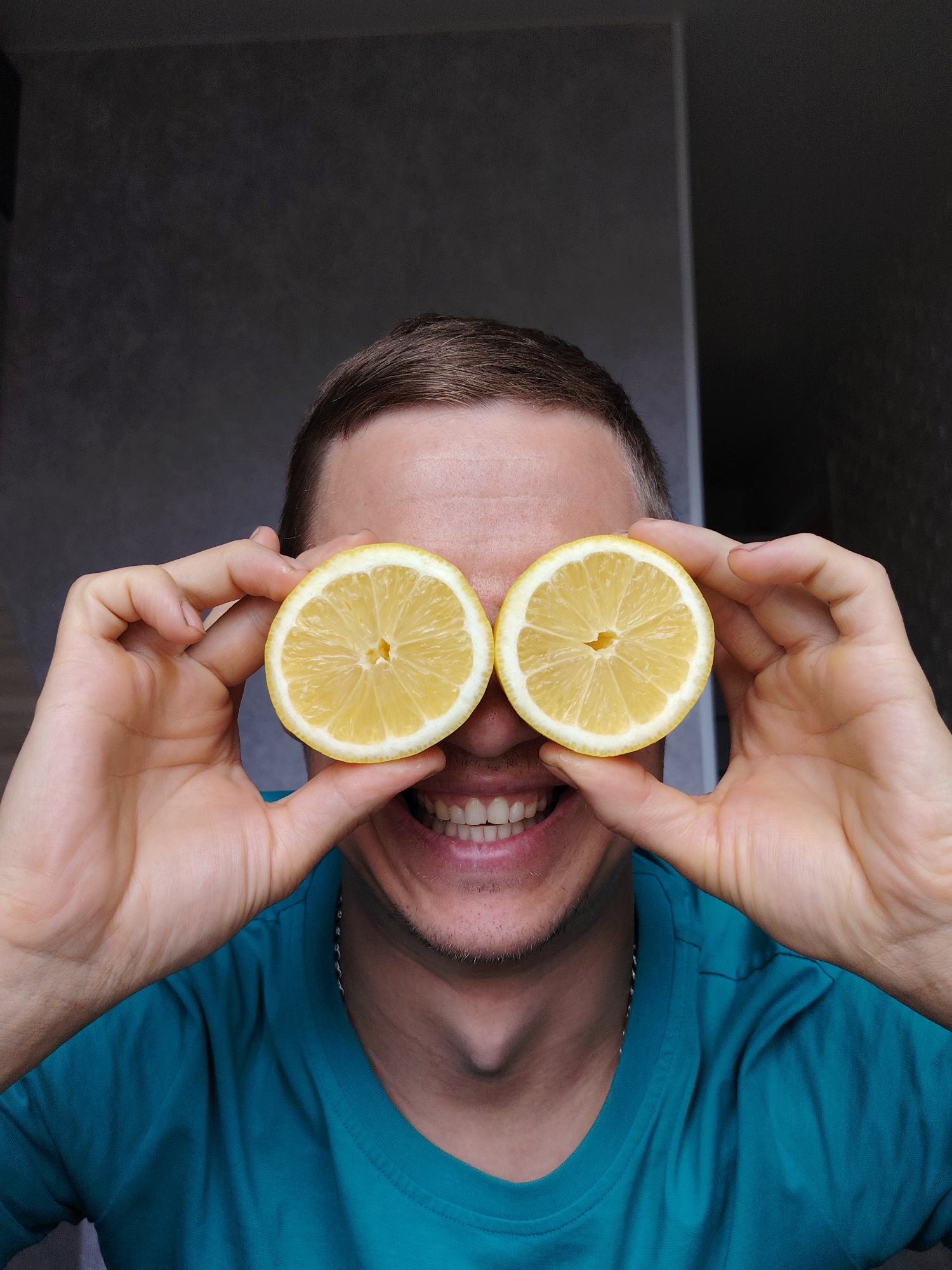 A close-up, eye-level portrait features a young adult male playfully posing indoors in Chaikovsky, Russia. He is positioned centrally, directly facing the camera. His face is largely obscured by two bright yellow lemon halves, which he holds over his eyes, creating a humorous "lemon-eye" effect. His hands are visible holding the lemons firmly. Below the lemons, his wide, toothy smile is clearly visible, conveying amusement and a cheerful mood.
The man has short, brown hair and a light complexion. He is wearing a vibrant turquoise t-shirt, and a thin, silver-colored chain is visible around his neck, resting on his shirt. The background is a plain, dark gray wall, suggesting an indoor setting with minimal distractions. A subtle vertical line is discernible on the right side of the background, possibly indicating a wall corner or door frame. The lighting is even, illuminating the subject without harsh shadows, and the overall impression is one of lightheartedness and fun. There is no visible text in the image.