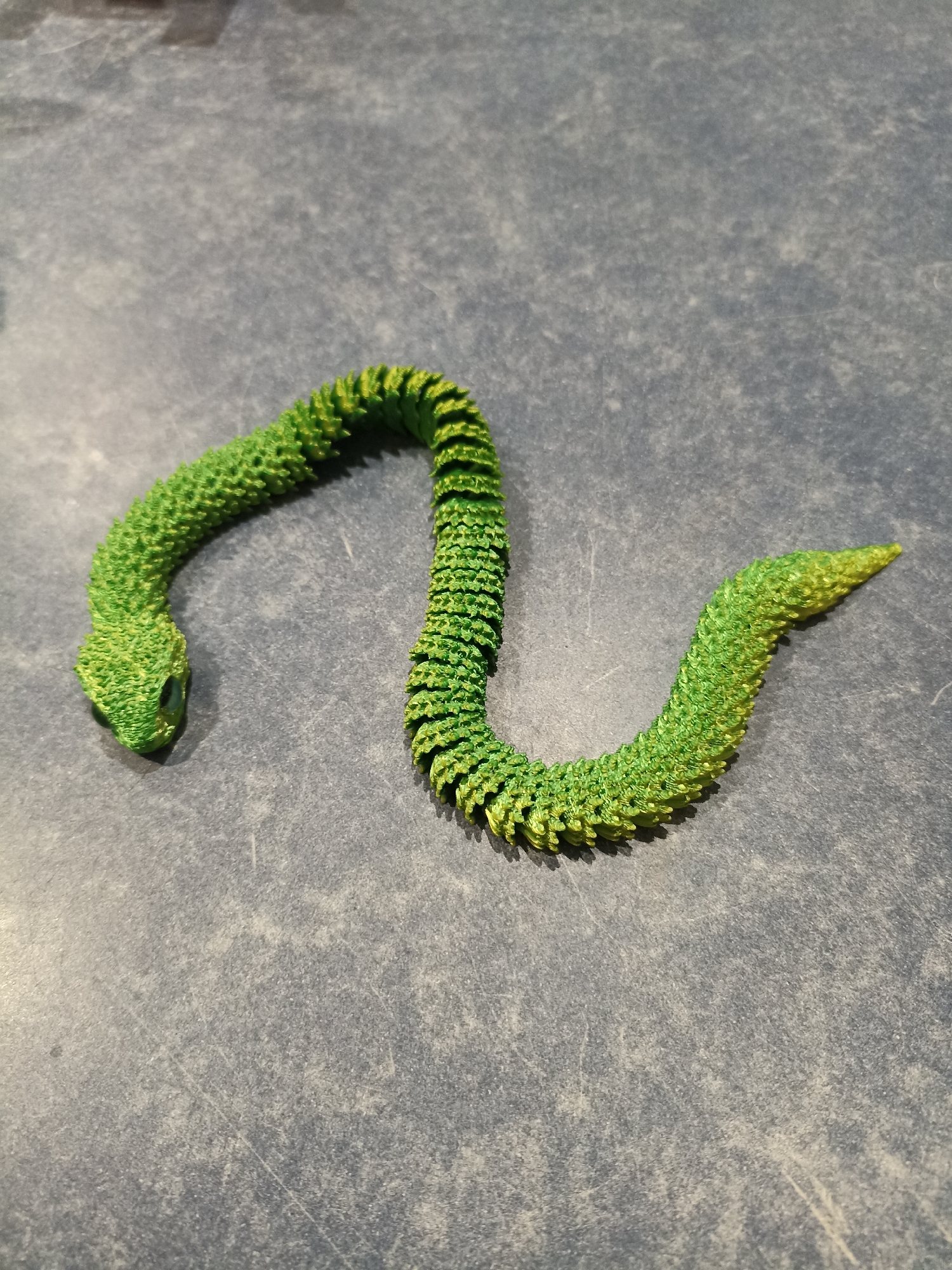 A green, segmented, snake-like toy is positioned on a dark, textured surface. The toy is coiled into a gentle S-shape, with its head on the left and tail on the right. The toy has visible green and yellow coloration, with a textured, scaled appearance. The surface appears to be a countertop or table with visible scratches and specks of light-colored debris. The lighting suggests an indoor environment, with a light source creating a subtle shadow to the right of the toy. The image is taken from a slightly elevated angle.