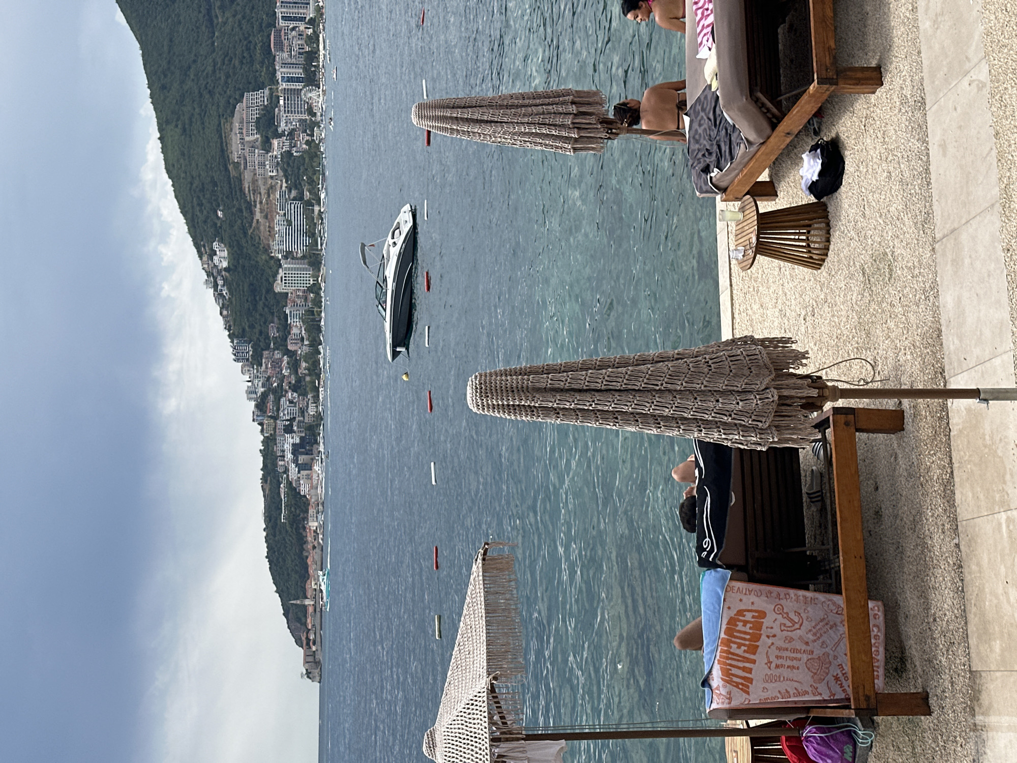 In Budva, Montenegro, this image depicts a cloudy day at a beachside resort. The foreground features two sun loungers with people relaxing. On the left, a person is lying on a lounger covered with a towel and what appears to be a blue cloth. To the right, two individuals are seated on a lounger, with one partially submerged in the water. Beach umbrellas provide shade over the loungers. In the middle ground, the clear blue water of the Adriatic Sea is visible, with several red and white buoys marking a swimming area. A sleek motorboat is anchored in the water. The background showcases the city of Budva, a picturesque coastal town with buildings cascading down a lush green hillside, dominated by a mountain peak. The overall scene conveys a sense of leisure and relaxation at a popular tourist destination.