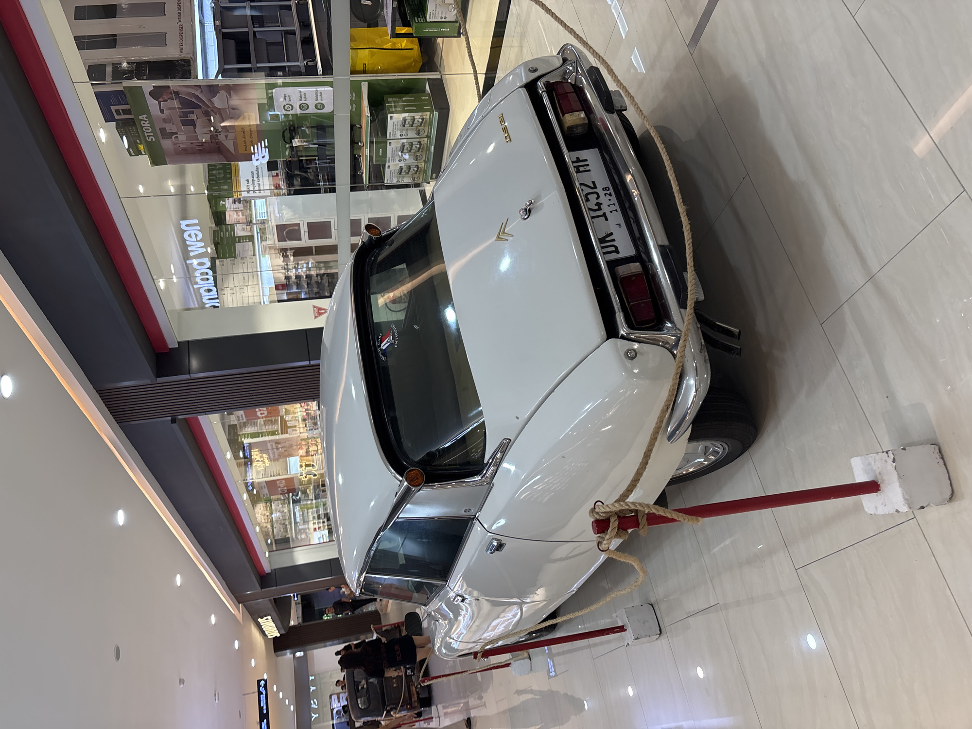 The image shows a white Citroën DS sedan on display in a brightly lit indoor location, likely a shopping mall. The car is roped off with red poles and heavy rope. Behind the car, shops can be seen. There are a few people visible in the background, near the car. The scene is in Kuta, Indonesia. The license plate on the car reads "DA 1452 AF," and it is valid until 11-28. A sticker on the car reads "ENTHUSIAS TOLIR SATUROMIS" and the word "kitral keb" appear to be below it.