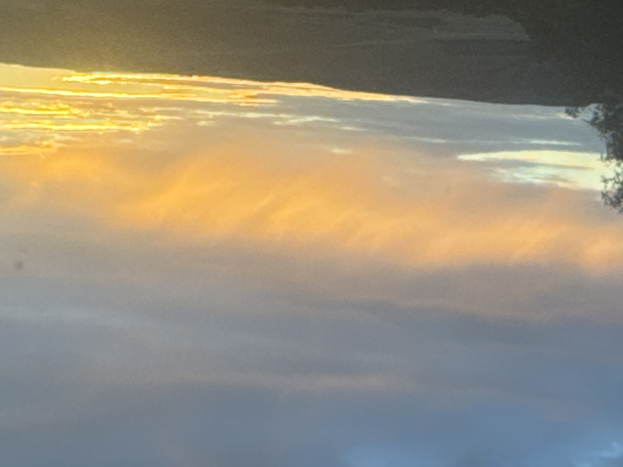 A landscape photograph captures a sunset over a hilly terrain. The sky is filled with clouds, transitioning from a deep blue at the top to vibrant orange and yellow hues near the horizon, indicating the setting sun. The lower portion of the image shows the silhouetted outline of a hill or mountain range. The foreground is dark, with a hint of vegetation visible on the left side. The scene is natural and peaceful, evoking a sense of twilight in the countryside. The image is undated and location specific to Vasil'evka, Russia.