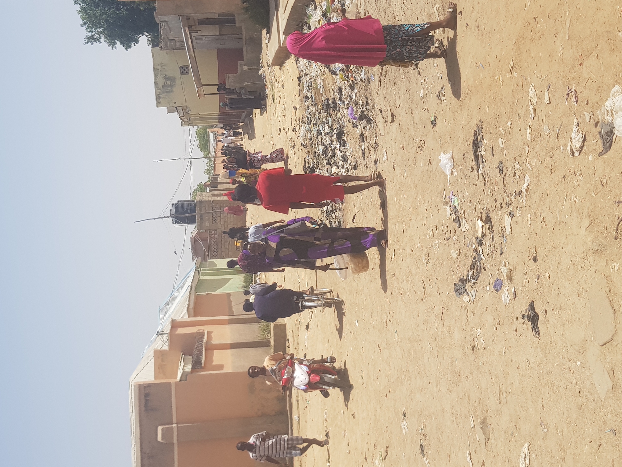 The image shows a dirt road in Dabawa, Nigeria, with buildings lining the side. There are people on the road, including individuals walking, riding bicycles, and riding a moped. Trash and debris are scattered along the road. Some of the people are wearing traditional clothing. A water tank is visible on top of one of the buildings. The sky is clear.