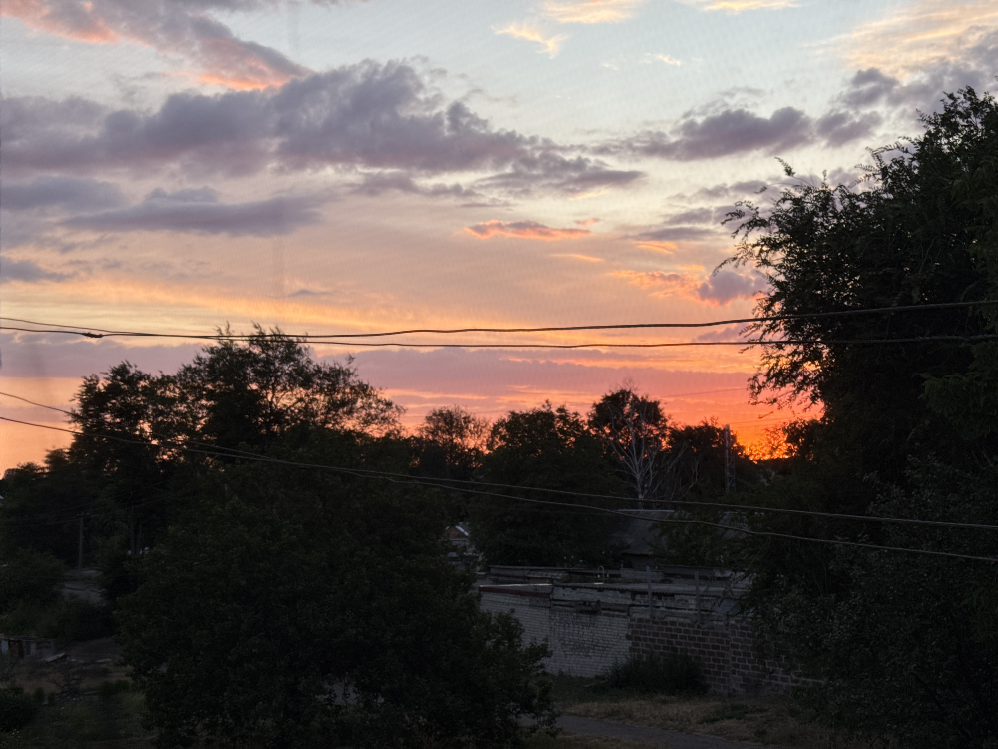 The image depicts a sunset over a semi-urban or rural area. The sky is a vibrant gradient of pink, orange, and pale blue, with scattered clouds. In the foreground, silhouetted against the colorful sky, are the dark shapes of trees and power lines. Below the sky, the landscape is dominated by dense foliage. A brick building with a weathered facade and metal elements is visible in the middle ground, along with glimpses of what appears to be a more sparsely developed area with some scattered structures. The overall impression is one of dusk in a tranquil, possibly suburban or countryside setting.