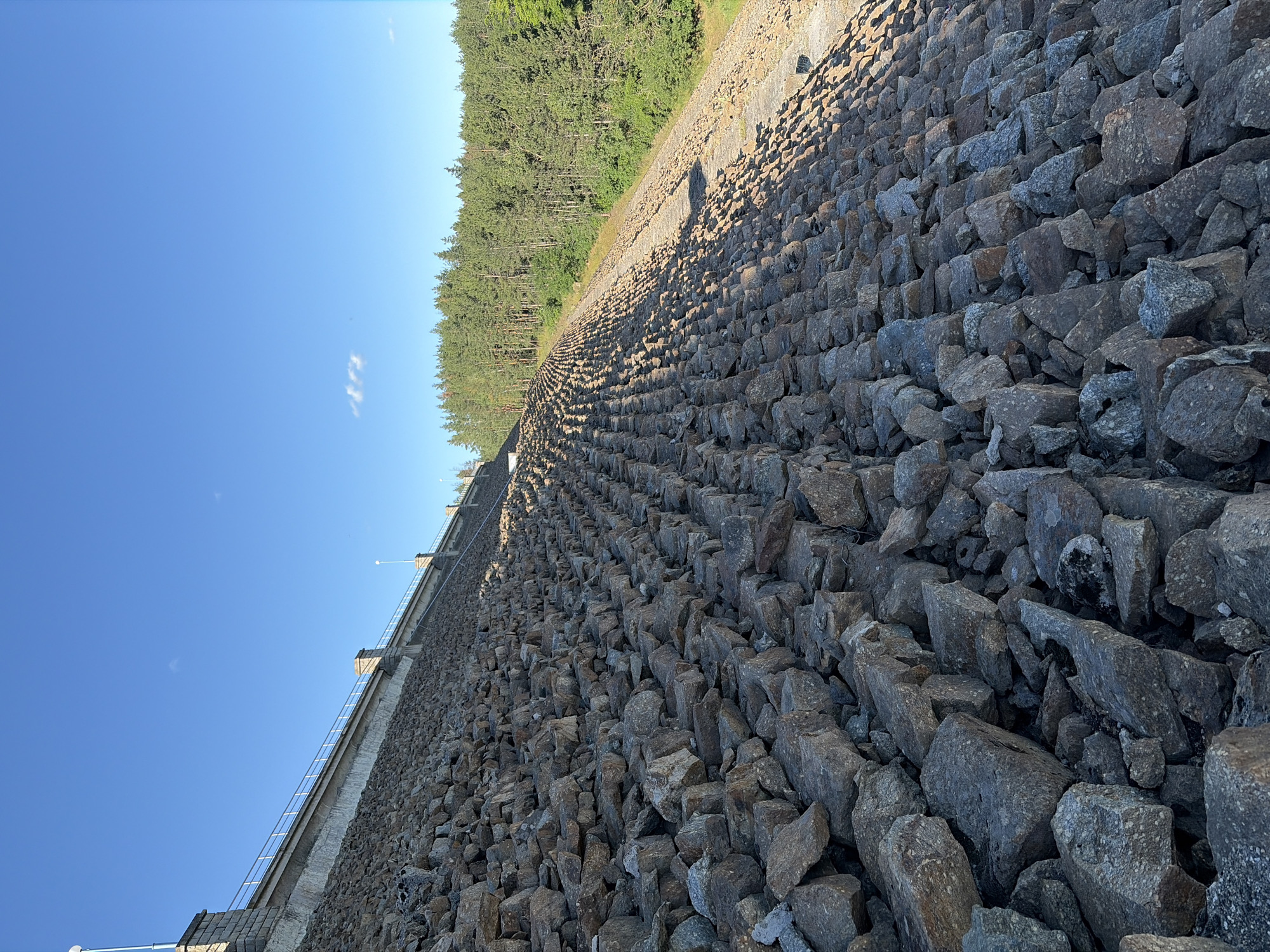 The image shows a close-up of the Koprinka Dam in Koprinka, Bulgaria. The dam is constructed of large rocks, creating a steep, textured slope. The rocks are varied in size and color, predominantly gray and brown, with some appearing to be granite. The surface of the dam is uneven and rugged, with shadows cast by the rocks indicating a bright, sunny day. At the top of the dam, a concrete structure with a metal railing is visible, suggesting a walkway or observation area. Beyond the dam, a dense forest of green pine trees stretches across the horizon under a clear blue sky with a few wispy white clouds. The overall scene is natural and man-made, showcasing the scale and material of the dam against a backdrop of lush vegetation.