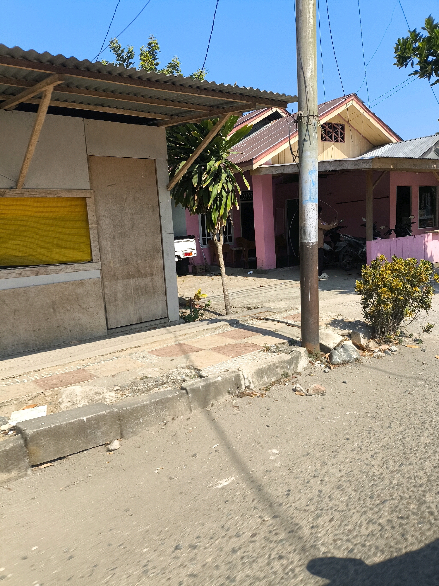 The image shows a street scene in Tilamuta, Indonesia. On the left is a small, unfinished building with a corrugated metal roof supported by wooden beams. It has a large wooden door and a rectangular window covered with yellow material. Adjacent to this building is a pink house with a brown and yellow roof. The house has an open carport with motorcycles parked underneath. A concrete utility pole stands in front of the pink house. A small yellow-leafed bush grows near the base of the utility pole. The street is paved with asphalt, and a concrete sidewalk with patterned tiles runs alongside it. The sky is clear and blue.