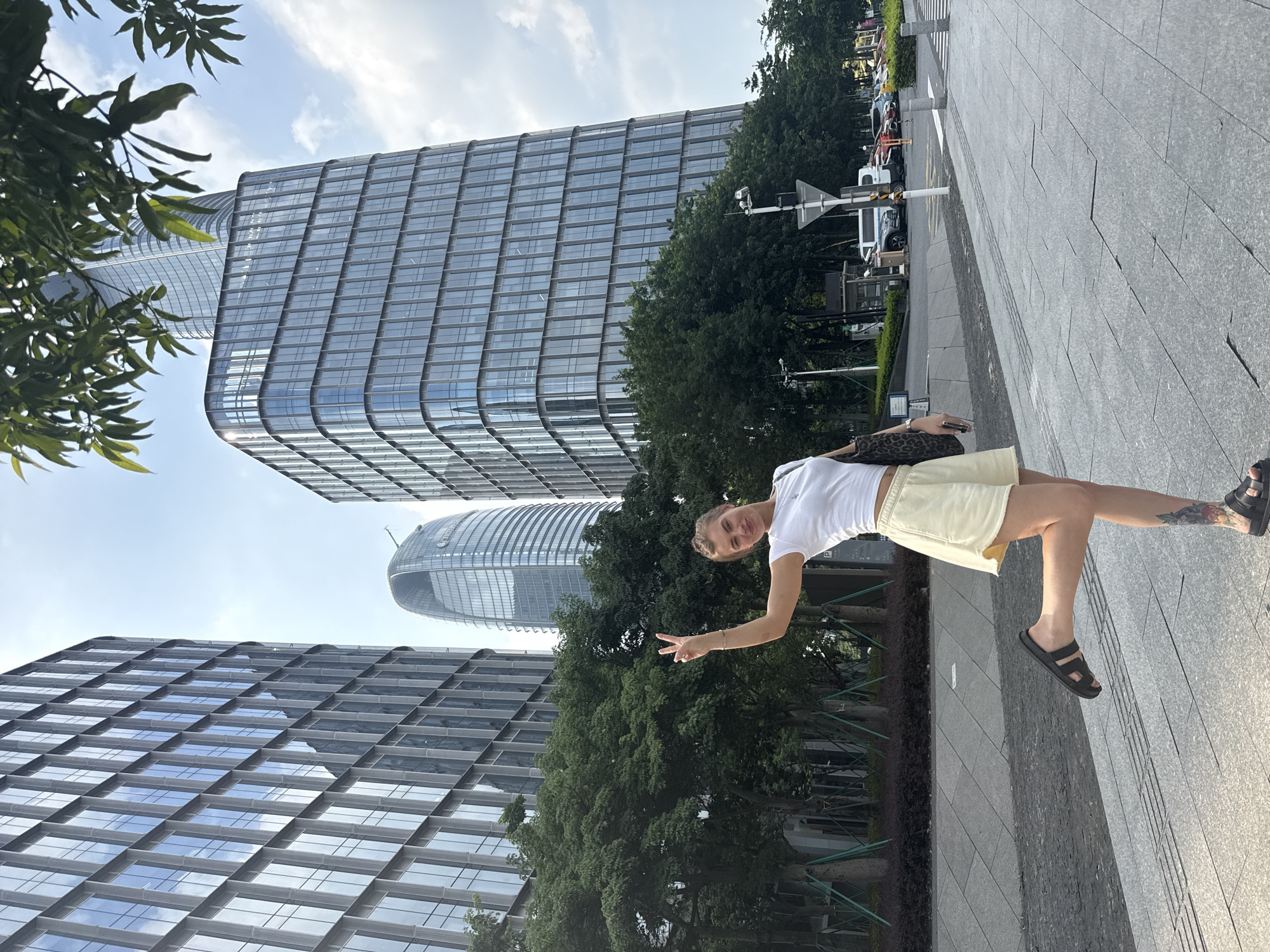 A young Caucasian woman with blonde hair pulled back stands on a paved sidewalk in front of a modern cityscape. She is wearing a white t-shirt and light yellow shorts, and she has black sandals on her feet. Her left arm is raised, and she is holding up two fingers in a peace sign. Her right hand is holding a large, leopard-print bag. She is looking towards the camera and smiling.
In the background, several tall, glass-fronted office buildings rise into a bright, partly cloudy sky. One building in the middle has a distinctive curved, egg-like shape. Lush green trees line the sidewalk in front of the buildings. The pavement is a light gray stone with a pattern of darker gray lines. Traffic can be seen in the distance, along with streetlights and signs. The overall impression is of a sunny day in a bustling urban environment. The scene appears to be located in the Haizhu District of China, characterized by its modern architecture and urban greenery.