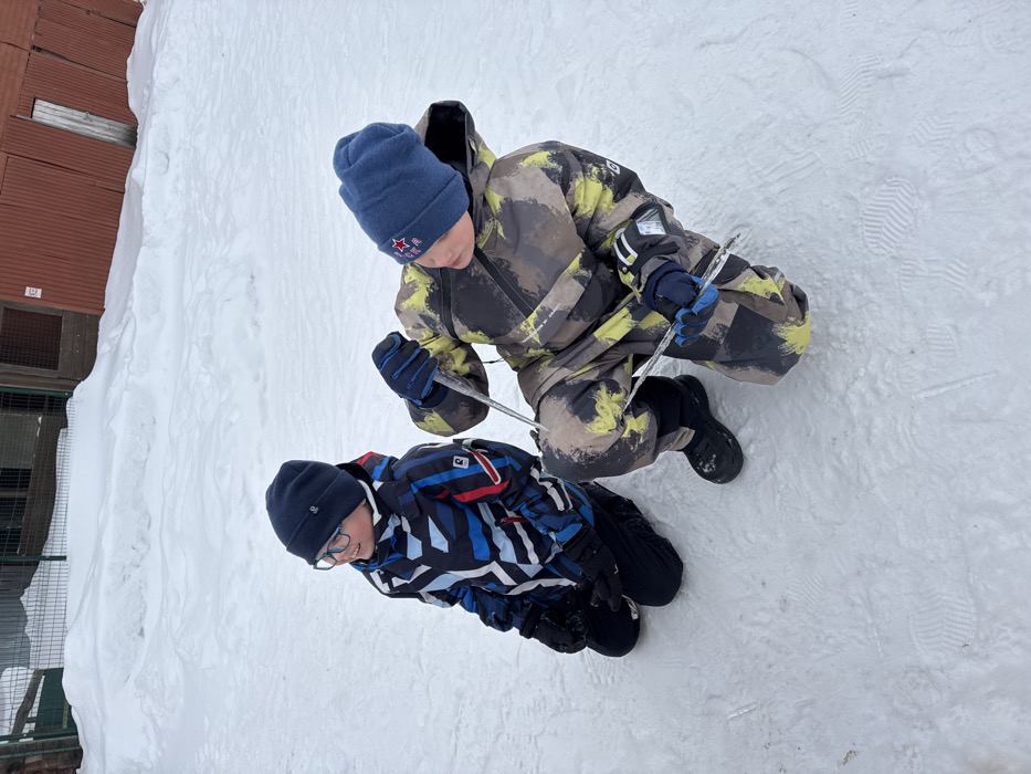 The image depicts an outdoor, snow-covered scene in Zaosinovo, Russia, featuring two young males, likely children, dressed in winter attire. The ground is uniformly covered in white snow with visible boot prints. In the background, a green metal fence or grate is partially obscured by snowdrifts on the left, while a reddish-brown wooden building with a vertical plank door and a dark numerical sign (possibly "30") is visible on the right.
On the left, one child is kneeling on the snow. He wears a dark blue knit beanie, blue-framed eyeglasses, a multi-colored winter jacket with blue, black, white, and red geometric patterns, black winter pants, black gloves, and black boots. His head is turned slightly to the right, and he exhibits a slight smile.
On the right, a second child is in a crouching or kneeling position. He wears a light blue knit beanie adorned with a red star emblem, a camouflage-patterned winter overall suit in shades of green, brown, black, and yellow, dark blue gloves, and black winter boots. He is actively holding two clear icicles: one long, vertical icicle is held by his right gloved hand, extending downwards, and a shorter, horizontal icicle is held by his left gloved hand near his left knee. He is looking downwards and to his left, seemingly focused on the icicles or the snow.