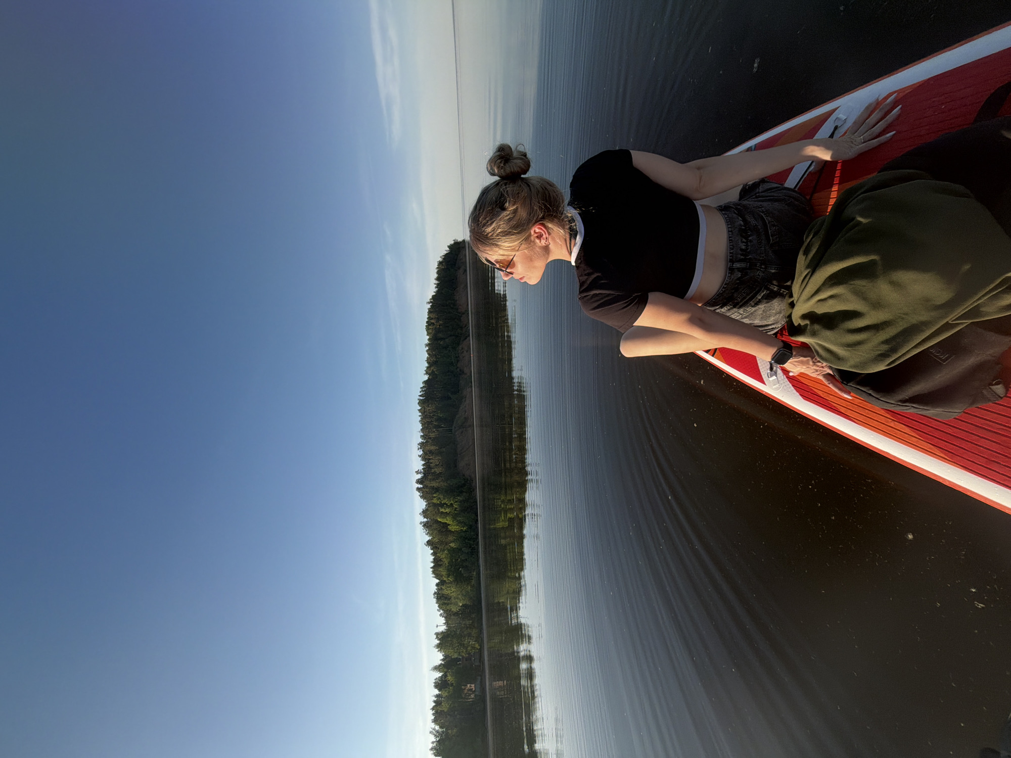 A woman lies on a red and white paddleboard on a body of water. She has her hair in a bun and wears sunglasses, a black crop top, and denim shorts. Part of a green jacket is on the board near her. The water is calm with reflections of a forested shoreline visible in the background. The sky is blue with some light clouds near the horizon. The image is taken at an angle.