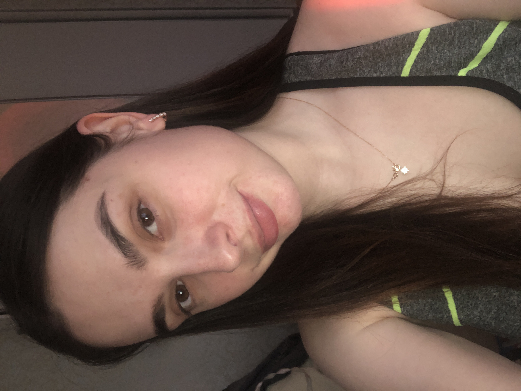 A portrait-oriented photo, rotated to show a young woman from an angled, slightly overhead perspective. She has long, dark brown hair, fair skin, and brown eyes. Her head is tilted, and she is looking directly at the camera with a gentle, closed-mouth smile, with a hint of her tongue visible. She is wearing a grey athletic top with bright neon green stripes. A delicate gold-colored chain necklace with a small, shiny charm featuring a clear stone rests on her chest. Her visible left ear features an ear cuff or multiple piercings adorned with a line of small, sparkling stones along the upper cartilage. The background is dark and out of focus, suggesting an indoor setting with dim, soft lighting. A reddish-orange glow is present in the upper right background, possibly from an artificial light source. There is no visible text in the image. The scene is located in Noviy, Russia, though the image itself does not contain specific visual cues to identify this location.