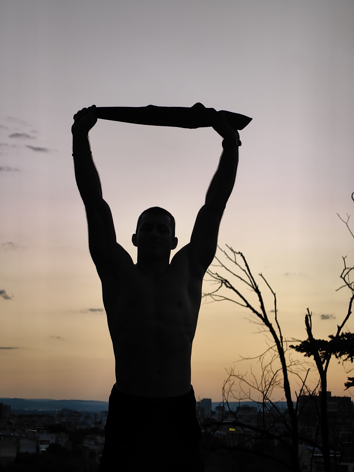 A silhouette of a shirtless man with dark hair stands with his arms raised above his head, holding a dark, elongated object horizontally. He is wearing dark shorts. He has a bracelet on his left wrist. The man is positioned in the foreground against a dusky sky with subtle clouds in shades of lavender and orange, indicative of sunset or sunrise. In the background, the silhouette of a city skyline with buildings is visible, suggesting an urban environment. Bare tree branches, also in silhouette, are present on the right side of the frame, partially obscuring the cityscape and sky. The overall scene evokes a sense of accomplishment or defiance. The city in the background is identified as Varna, Bulgaria.
