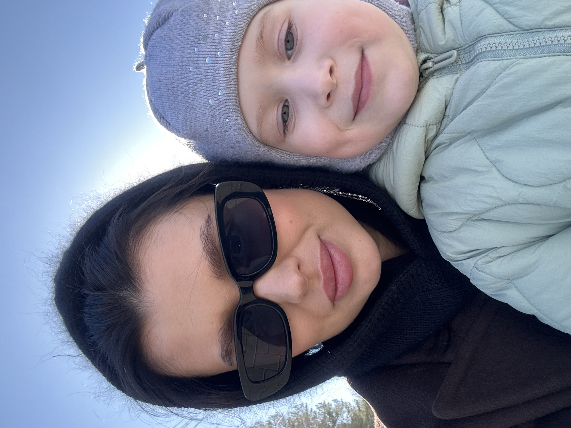 A woman wearing sunglasses and a hooded brown coat takes a selfie with a young child wearing a grey hat and light green puffy jacket. The background is a bright blue sky and sunlight. Part of the scene at the bottom shows some trees. This picture could have been taken in Chaikovsky, Russia.